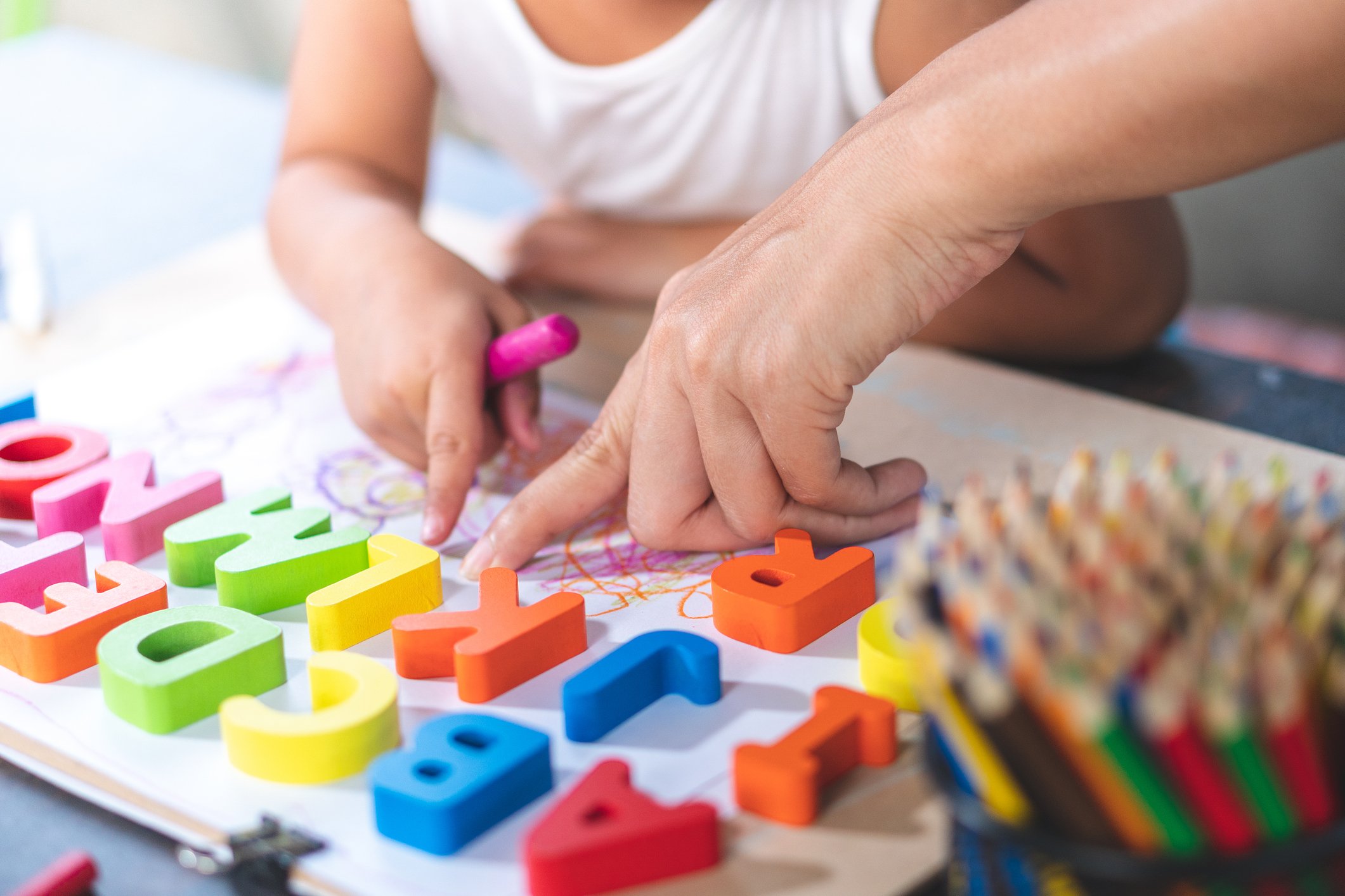 Close-up of a child's hand and an adult's hand playing with colorful foam alphabet letters on a table, with a container of colored pencils nearby.