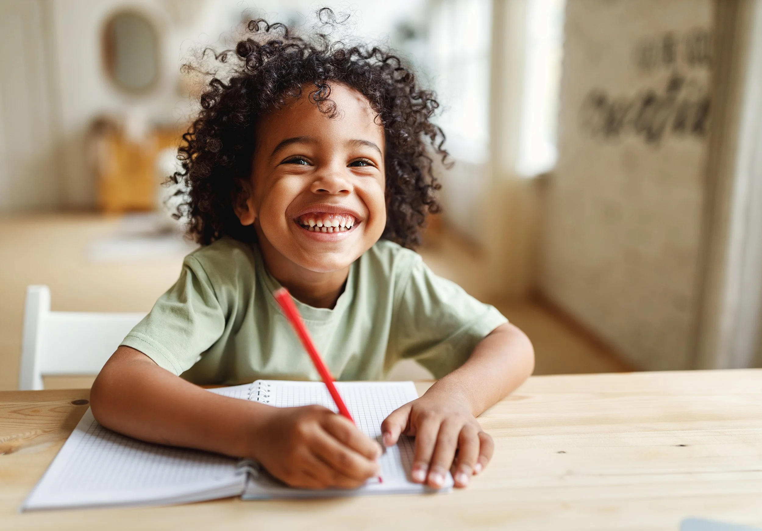 A smiling young child with curly hair sitting at a wooden table, writing in a notebook with a red pencil.