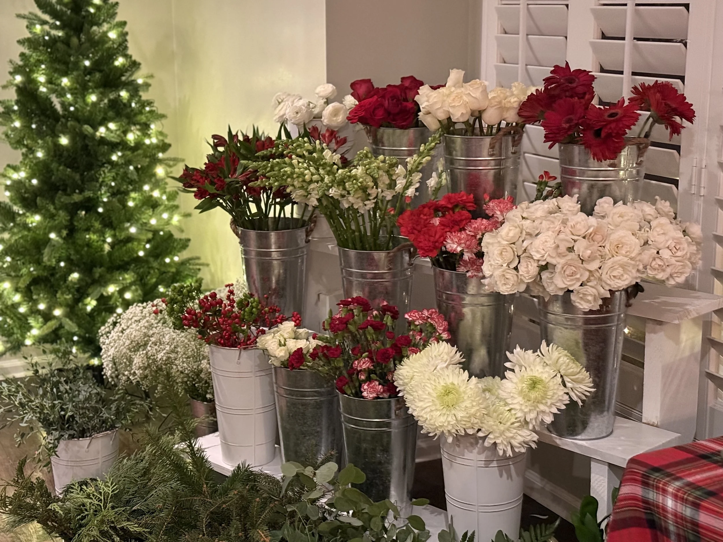 Display of various flowers in metal and white containers with a decorated Christmas tree to the left in a cozy room.