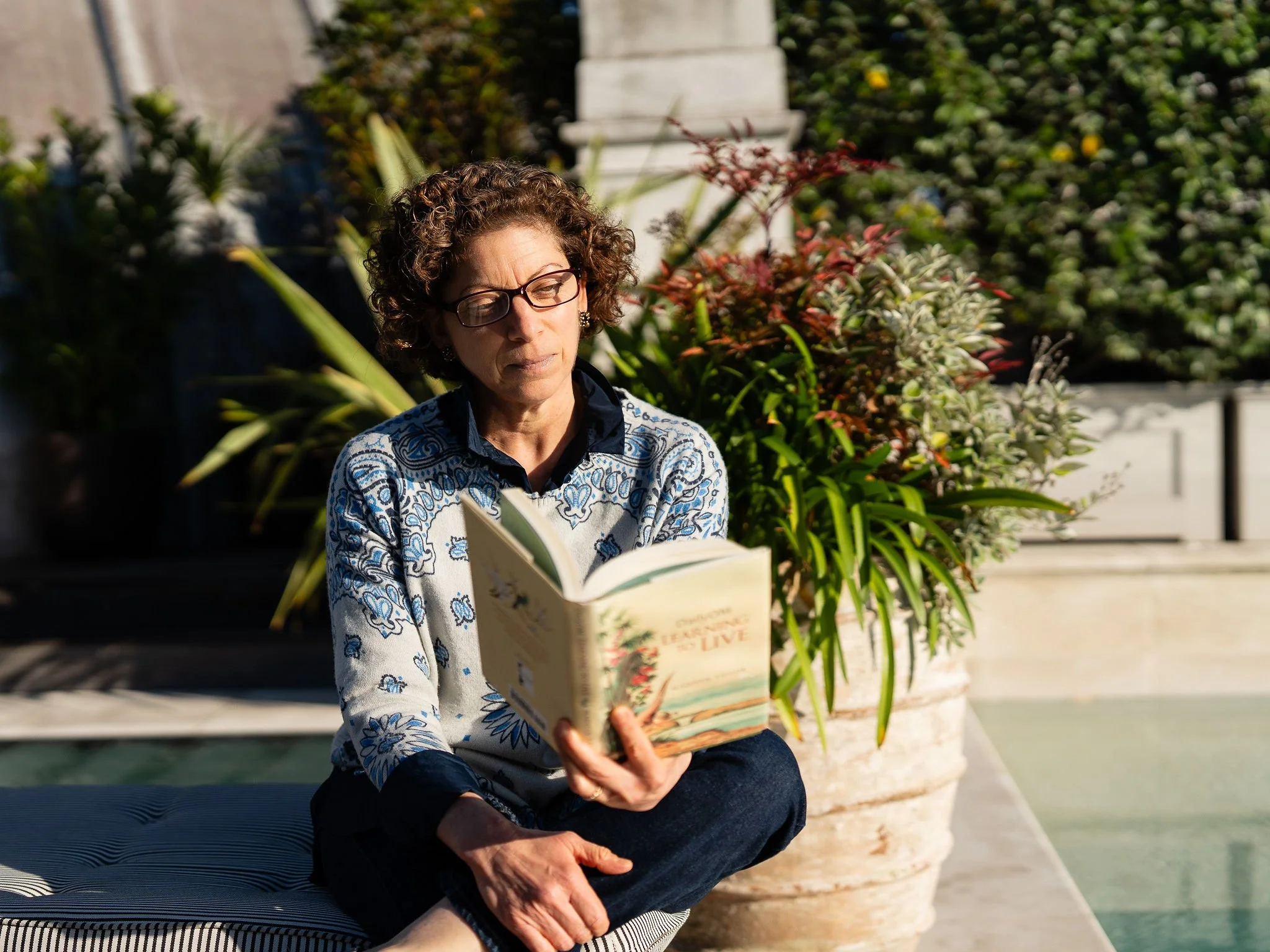 A woman with curly hair and glasses sitting outdoors on a striped cushion, reading a book, with large potted plants and greenery in the background.