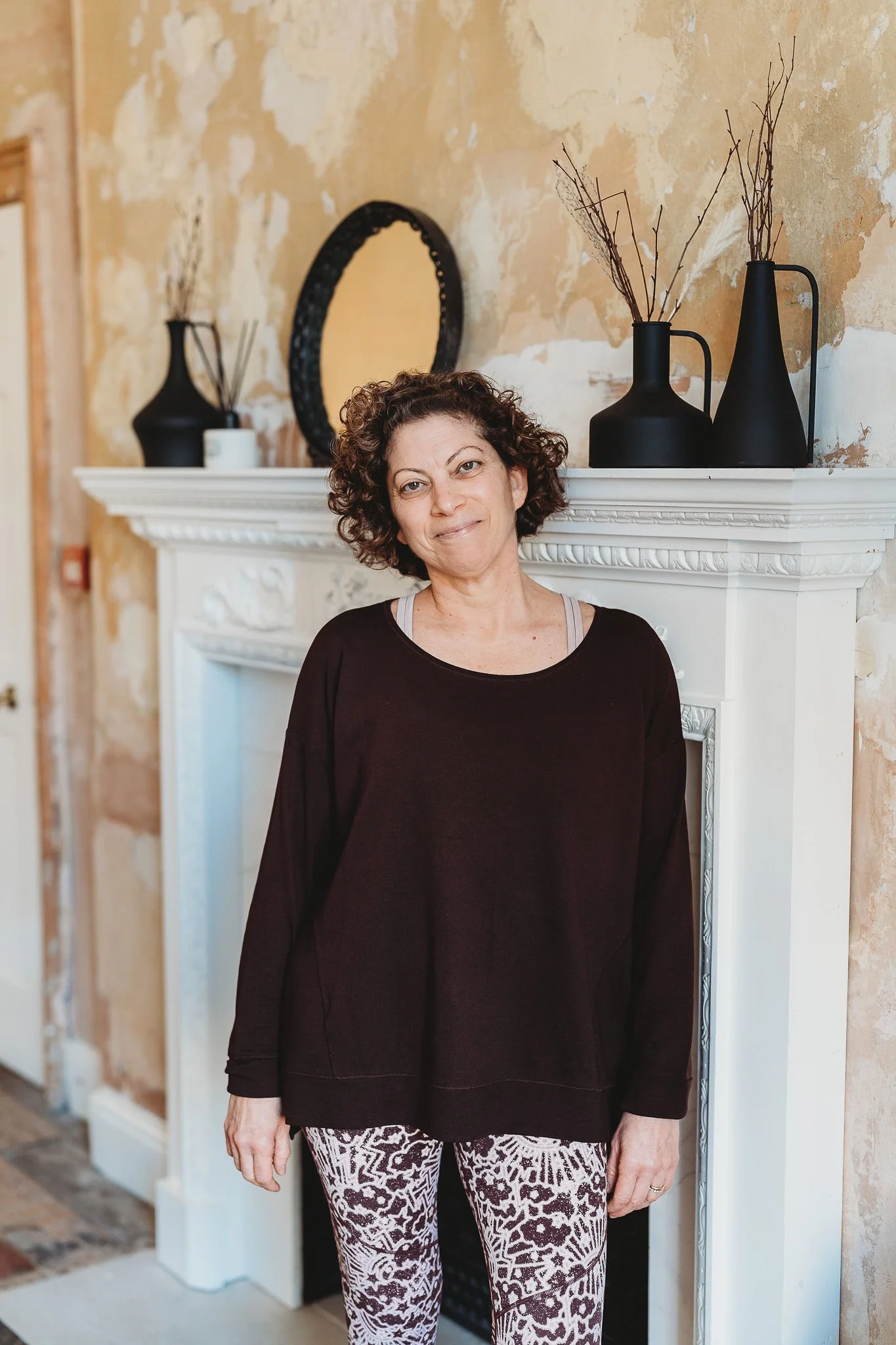 A woman with curly hair and a black long-sleeve shirt standing in front of a white fireplace with black vases and decorative items on top and a mirror on the wall behind.