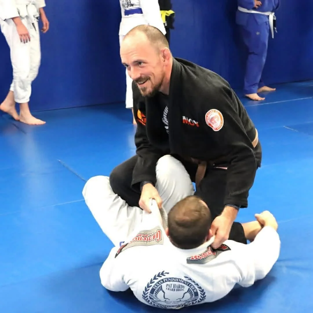 Two martial artists practicing Brazilian Jiu-Jitsu on a blue mat with a man in a black gi smiling and holding a smaller man in a white gi in a grappling position, surrounded by other practitioners in gis in the background.