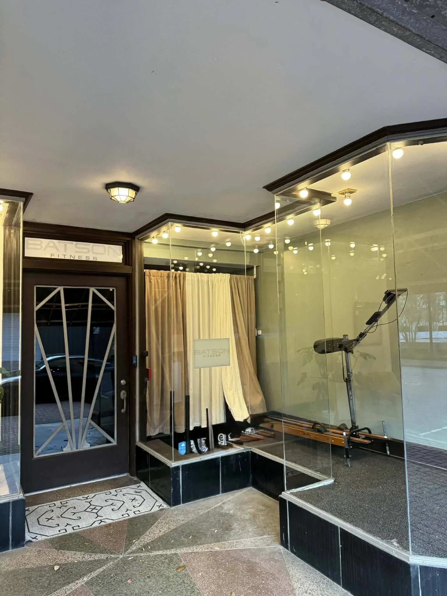 View of the entrance to a fitness facility called BATS ON FITNESS, with a glass wall revealing exercise equipment inside, including a rowing machine.