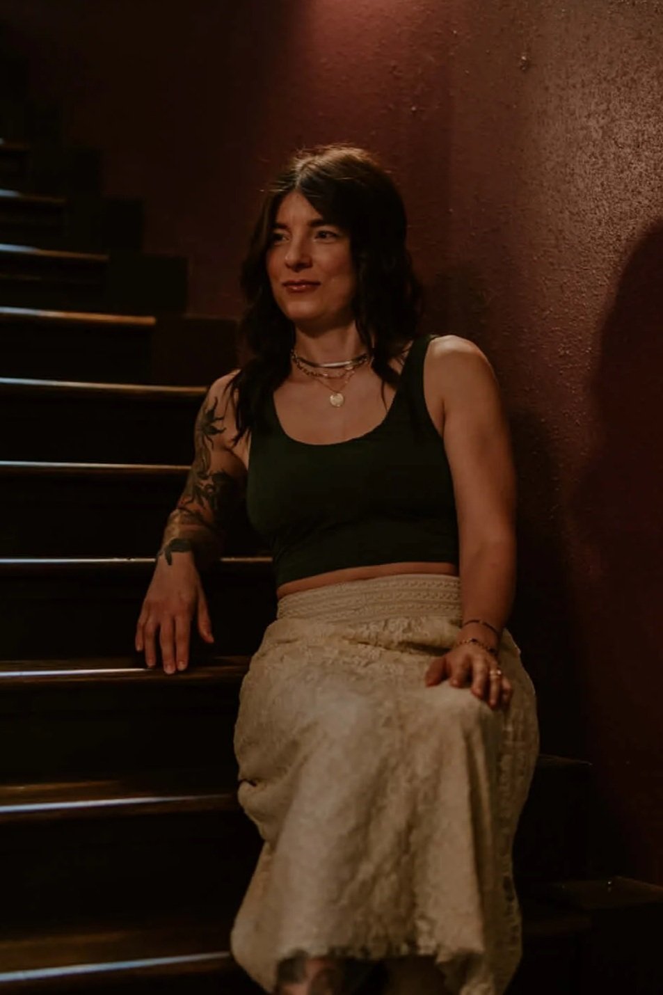 A woman with dark hair and tattoos on her arm, sitting on stairs next to a textured red wall, wearing a black crop top and a long beige skirt, accessorized with necklaces and bracelets, in a dimly lit setting.