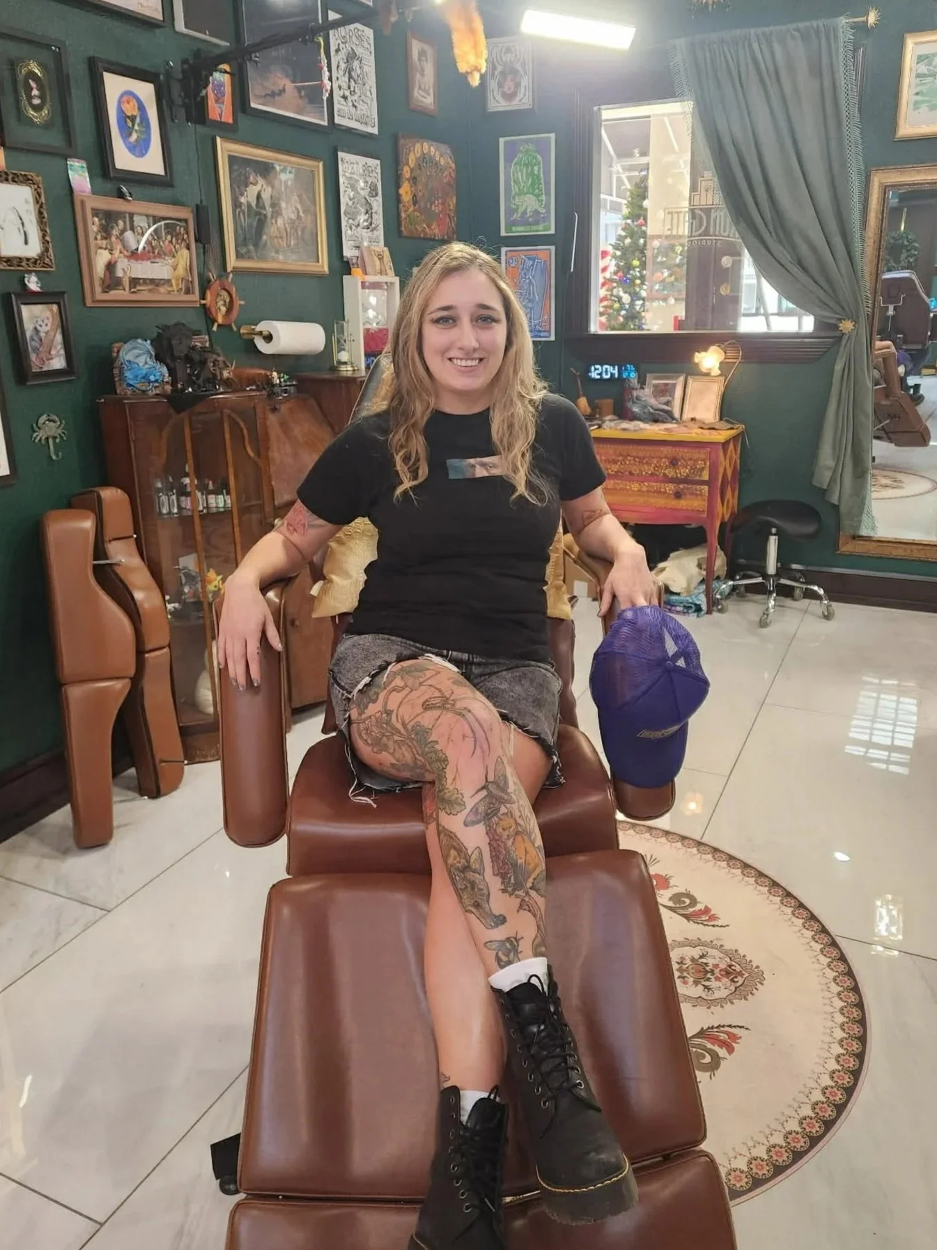 A young woman with blonde hair and tattoos on her legs is seated on a vintage barber chair in a tattoo studio. She is smiling, wearing a black t-shirt, denim shorts, and black boots. There is a blue cap hanging from her arm.