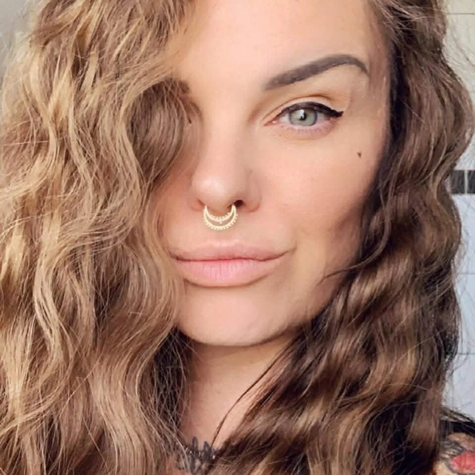 Close-up photo of a woman with light skin, wavy brown hair, and green eyes, wearing a septum ring and eyeliner.