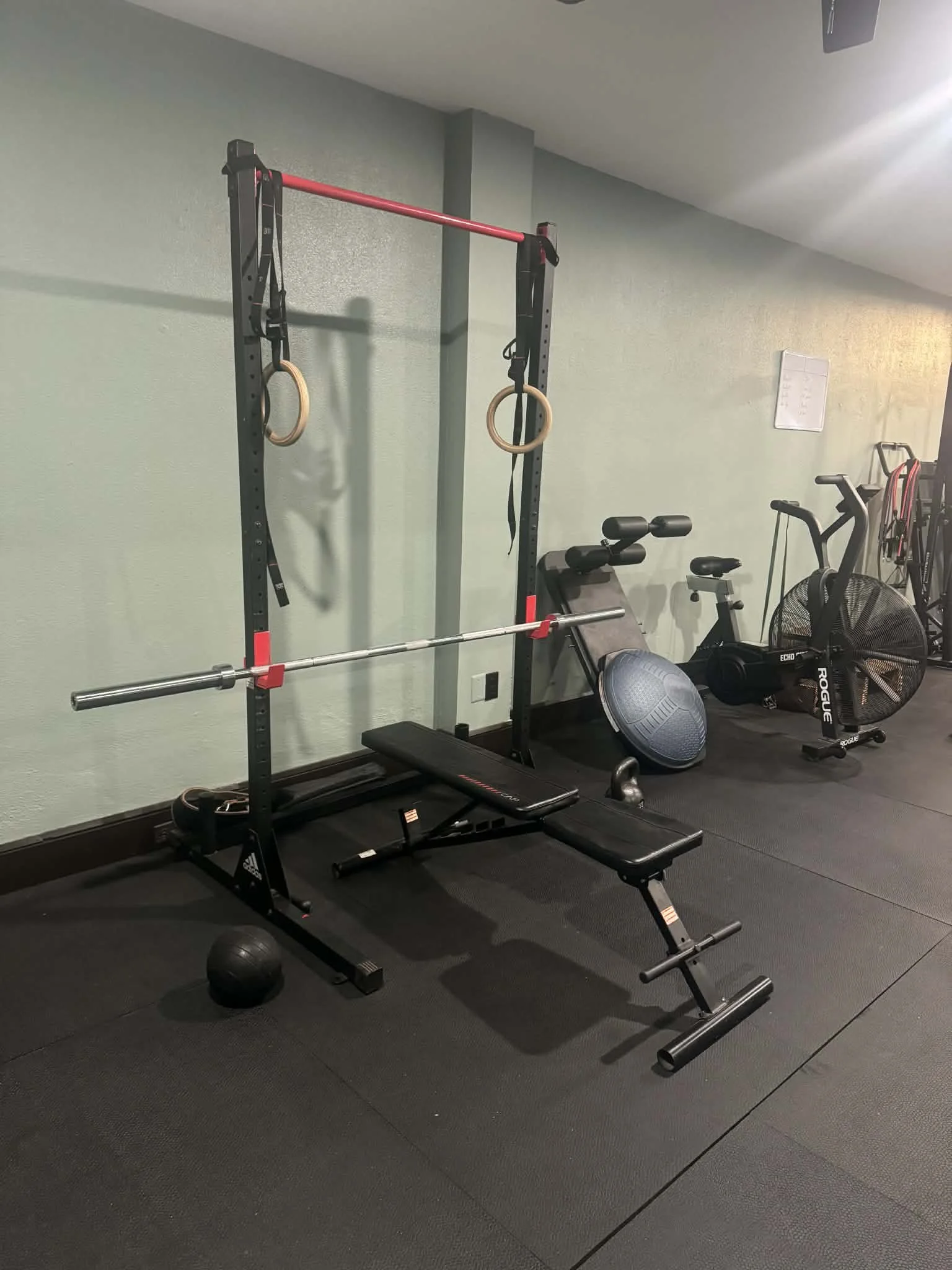 Home gym with a squat rack, gymnastic rings, a flat weight bench, a medicine ball, a stability ball, and a fan on black rubber flooring.