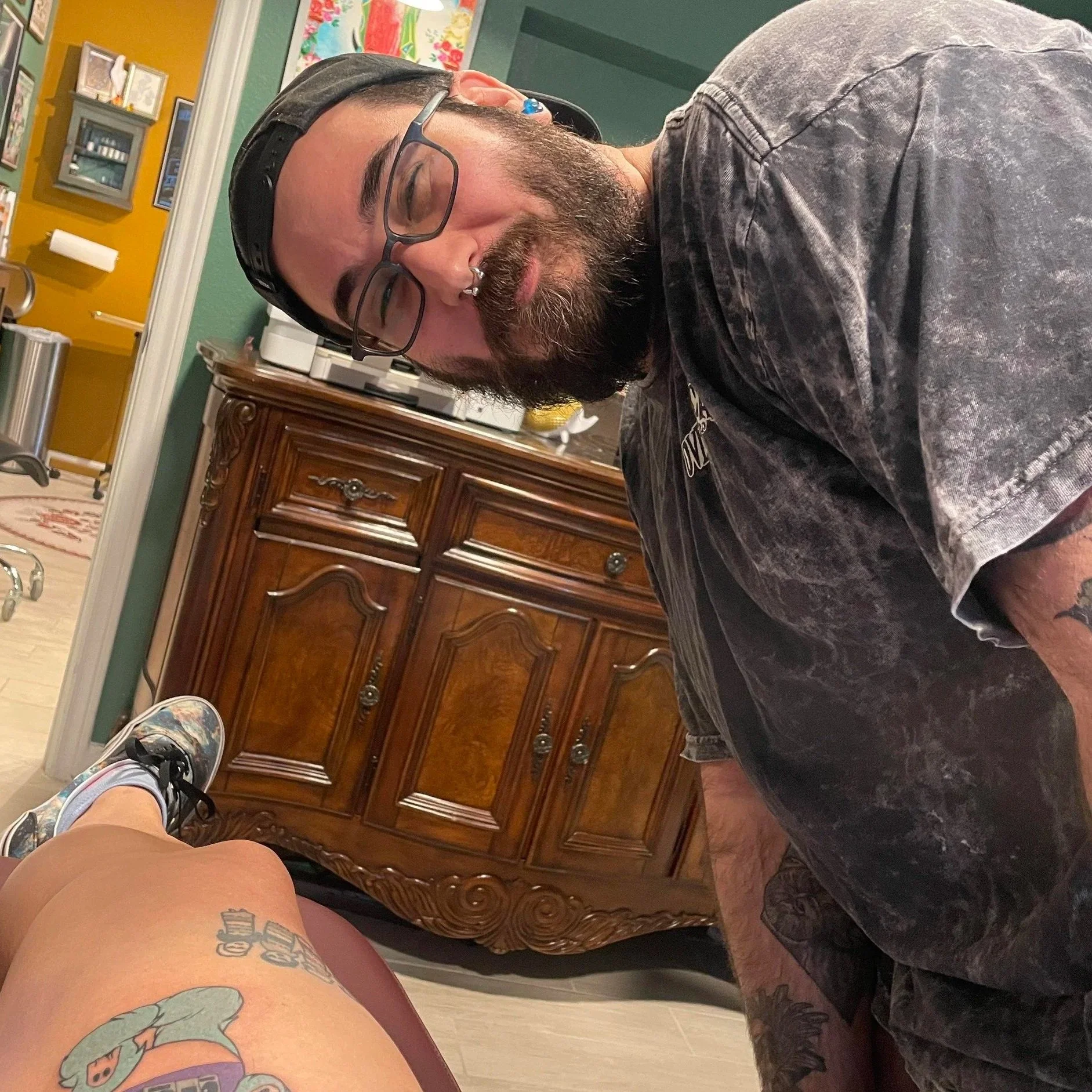 A man with glasses, a beard, and a nose ring smiling at the camera, leaning over a tattooed person's leg. The setting appears to be a tattoo parlor with wooden furniture and colorful artwork on the wall.