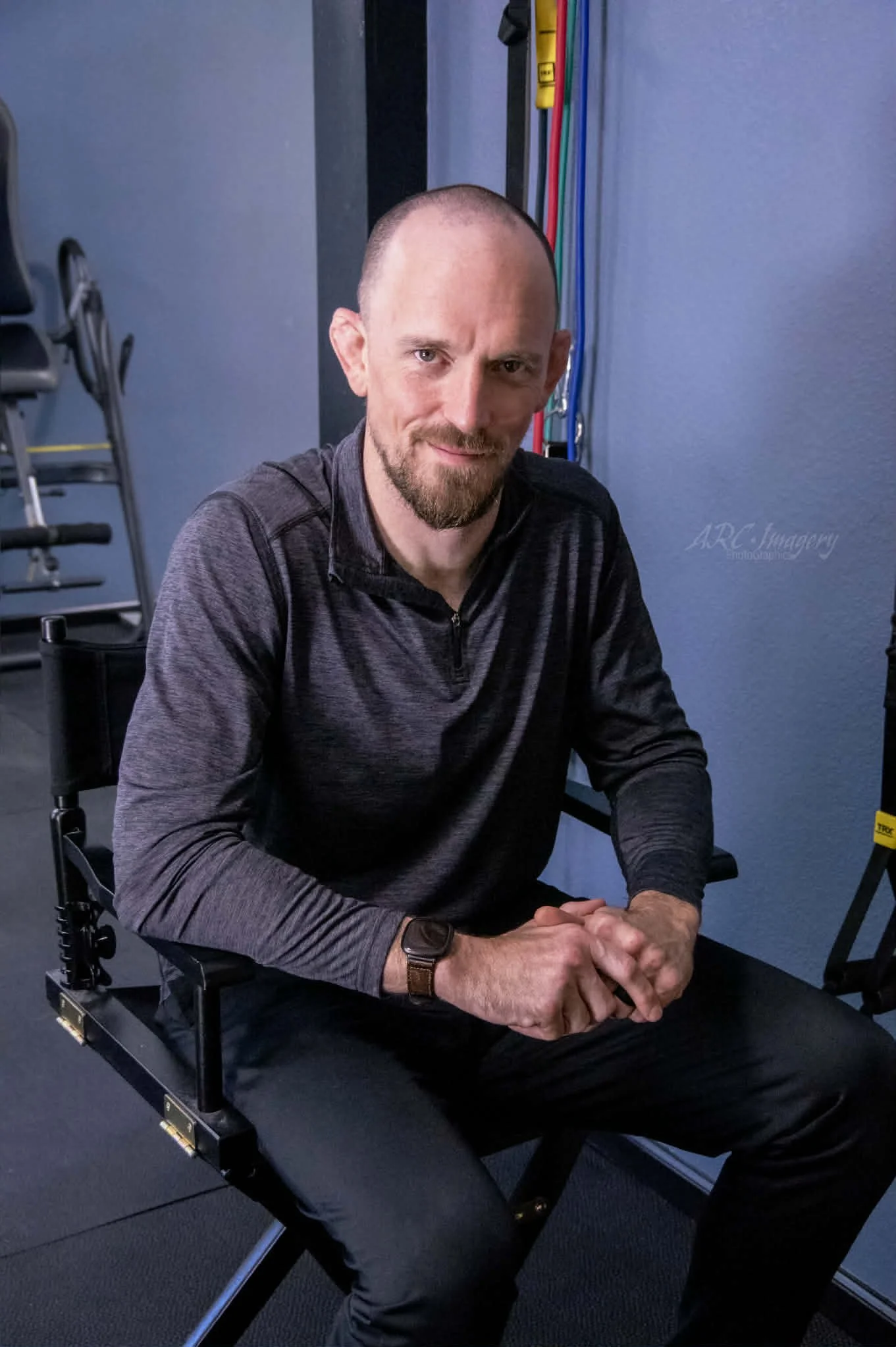 A man with a bald head, beard, and mustache sitting in a wheelchair, wearing a dark long-sleeve shirt, in a room with blue walls, exercise equipment, and colorful cables in the background.