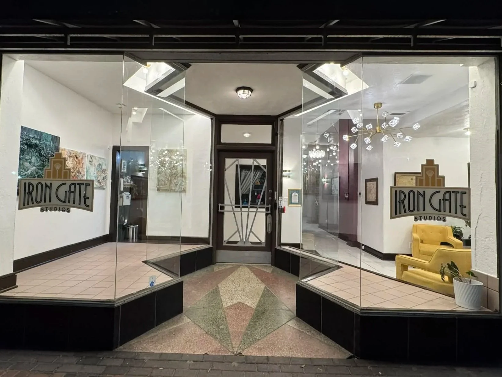 The window display of Iron Gate Studios shows the interior with artwork on the walls, several chandeliers, a yellow armchair, and a potted plant. The studio's logo is on the glass, featuring a stylized building.