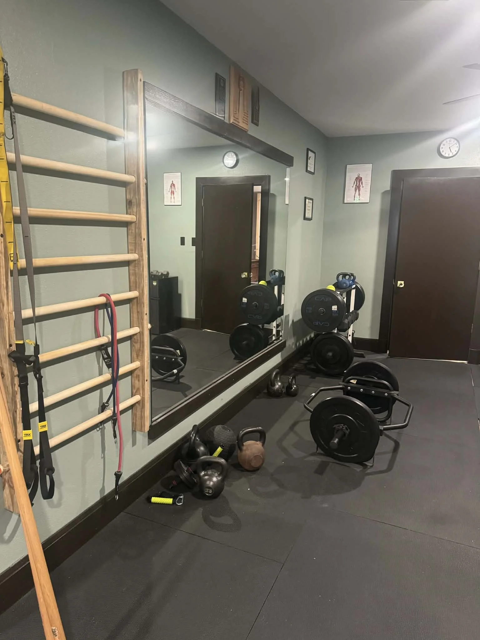Home gym with black rubber flooring and various workout equipment, including kettlebells, dumbbells, weight plates, a mirror, and a wall-mounted wooden ladder for stretching or resistance exercises.
