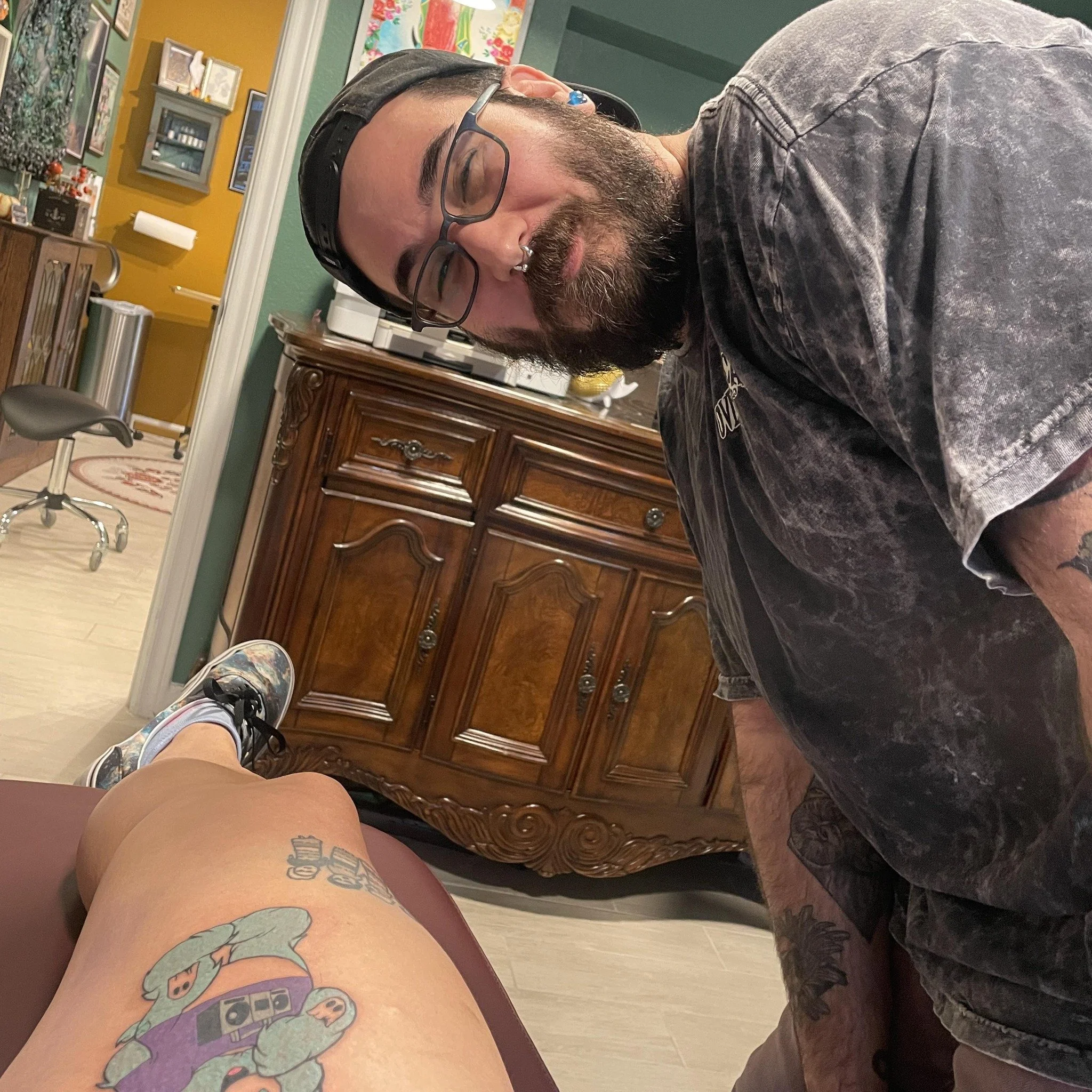 A man with a beard, glasses, and a nose ring looks down at a person's leg with a tattoo of a dragon, a cassette tape, and a cartoon character. The man is wearing a black cap and a dark, faded t-shirt. The background shows a wooden cabinet, colorful artwork, and a desk with various items in a room with yellow walls.