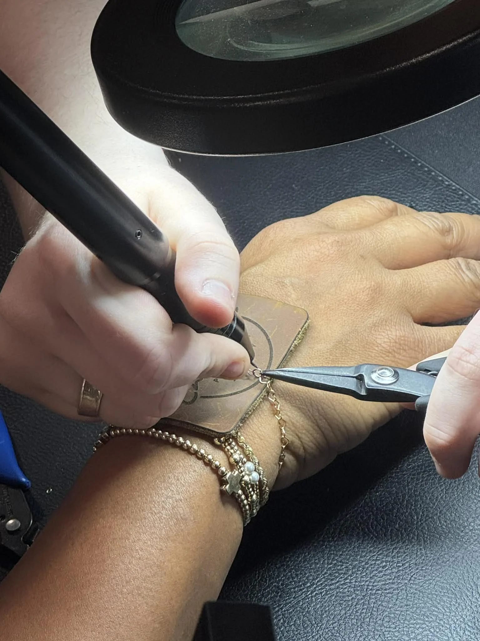 A person is engraving a design onto a small metal piece using a rotary tool. The person's hand is on a black textured surface, and they are wearing multiple bracelets on their wrist.