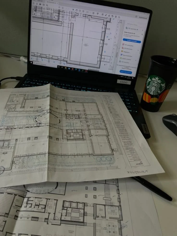 Draft architectural blueprints on a desk, a laptop showing a digital building plan, a Starbucks cup, and a black pen.