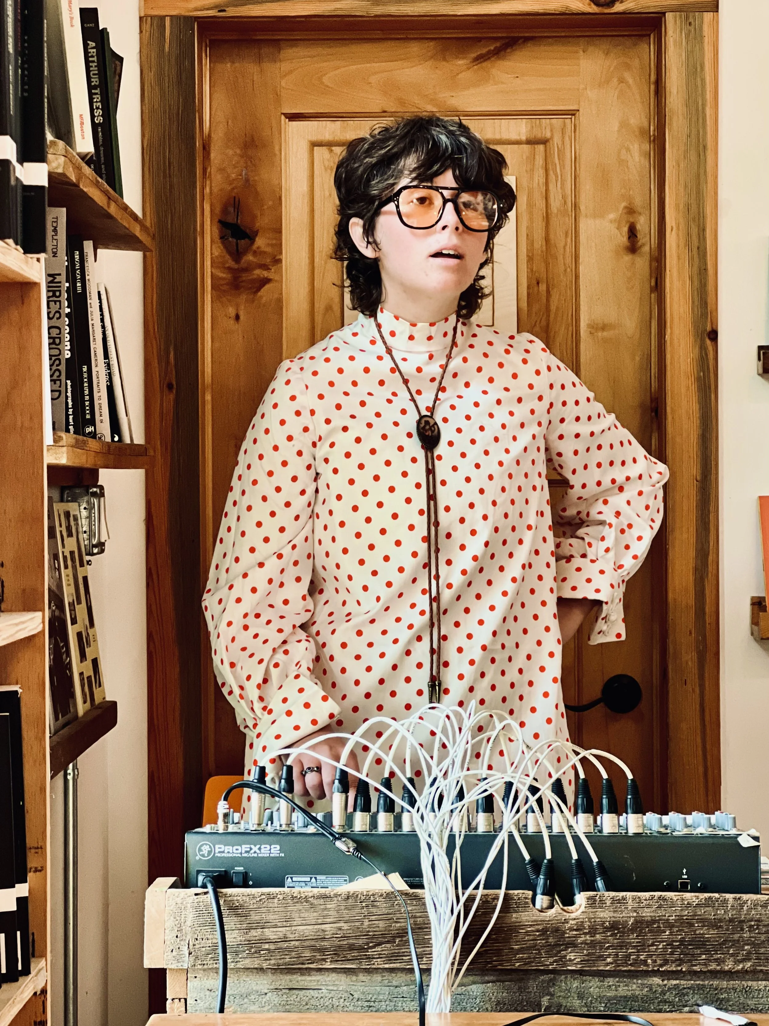 Non-binary human wearing red polka dot long sleeved outfit and bolo tie looks off to the right in creative exploration