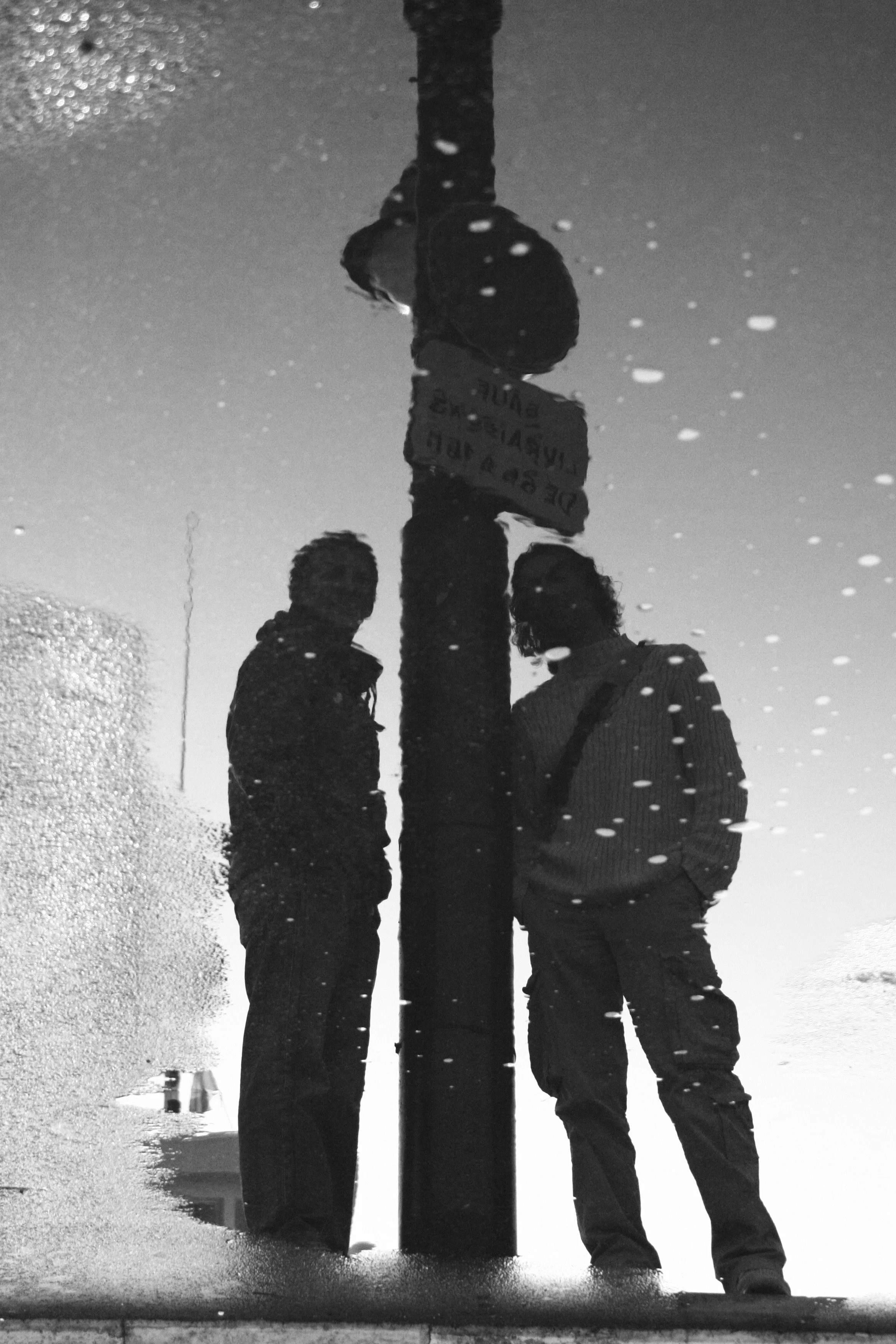 Reflection of two people standing next to a street sign in a puddle on a rainy day.