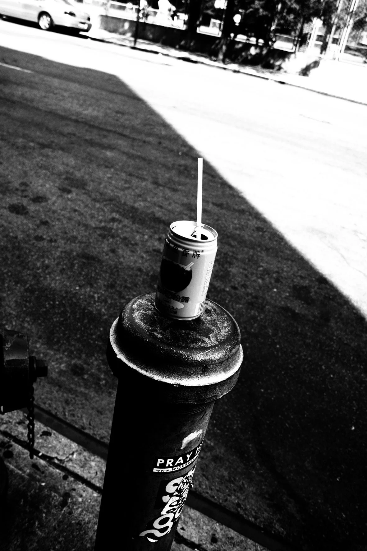 A soda can with a straw balanced on a fire hydrant on the sidewalk on a sunny day, with street and buildings in the background.