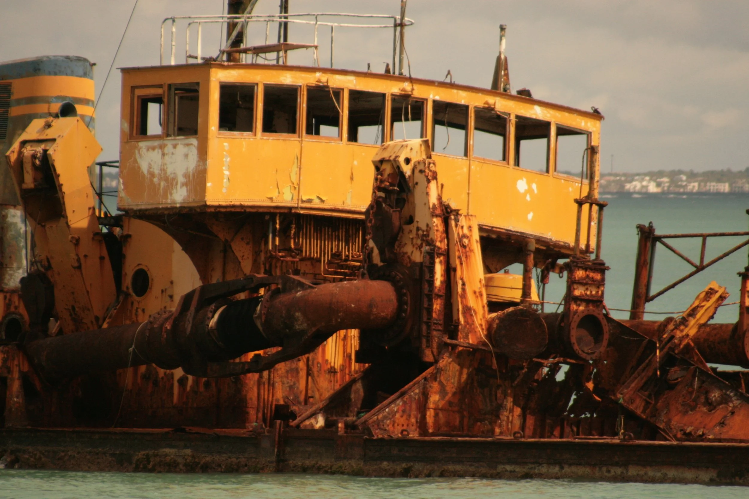 A rusty, abandoned ship in water, with a yellow superstructure, and the background shows distant land and buildings.