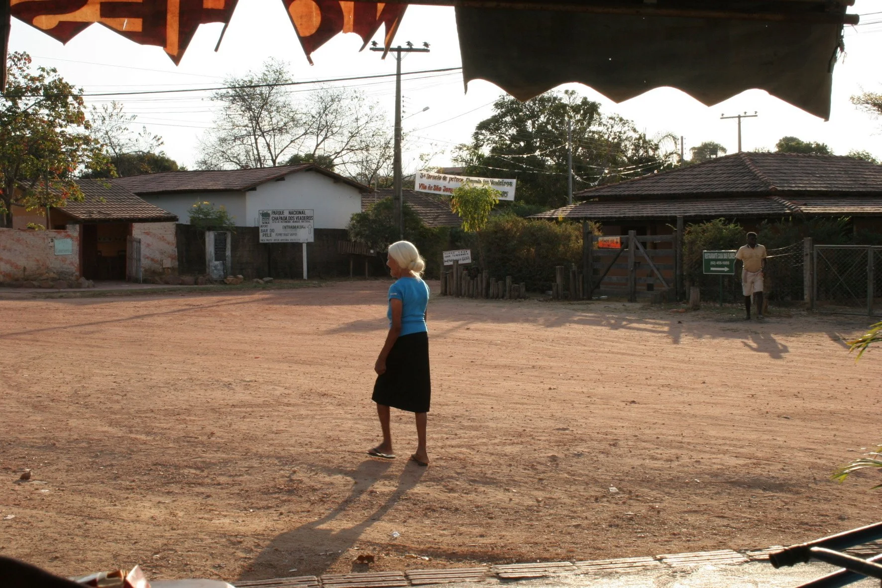 A rural village scene with two women walking on a dirt street in the late afternoon, surrounded by small houses and trees.