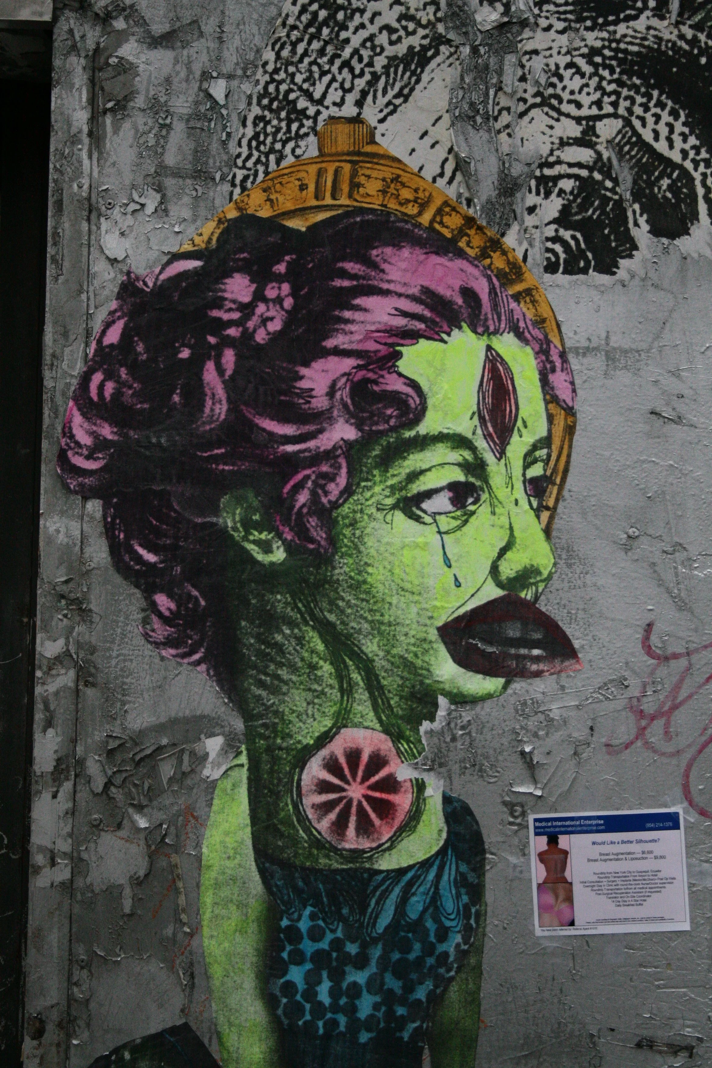 Street art of a surreal female figure with green skin, purple hair, and a third eye on her forehead, depicted with a tear, wearing a blue top with black dots, against a textured, peeling wall.