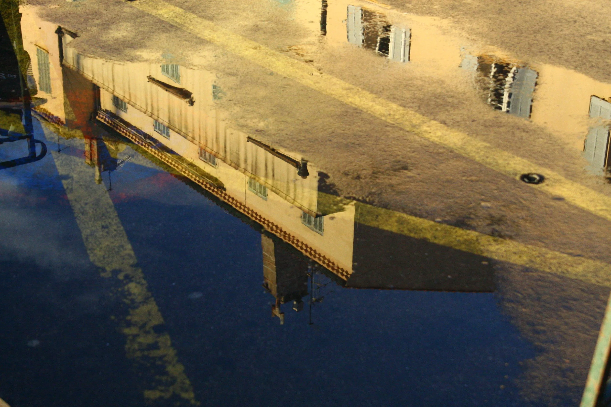 Reflection of a yellow building with windows and a chimney in a puddle of water on the ground.