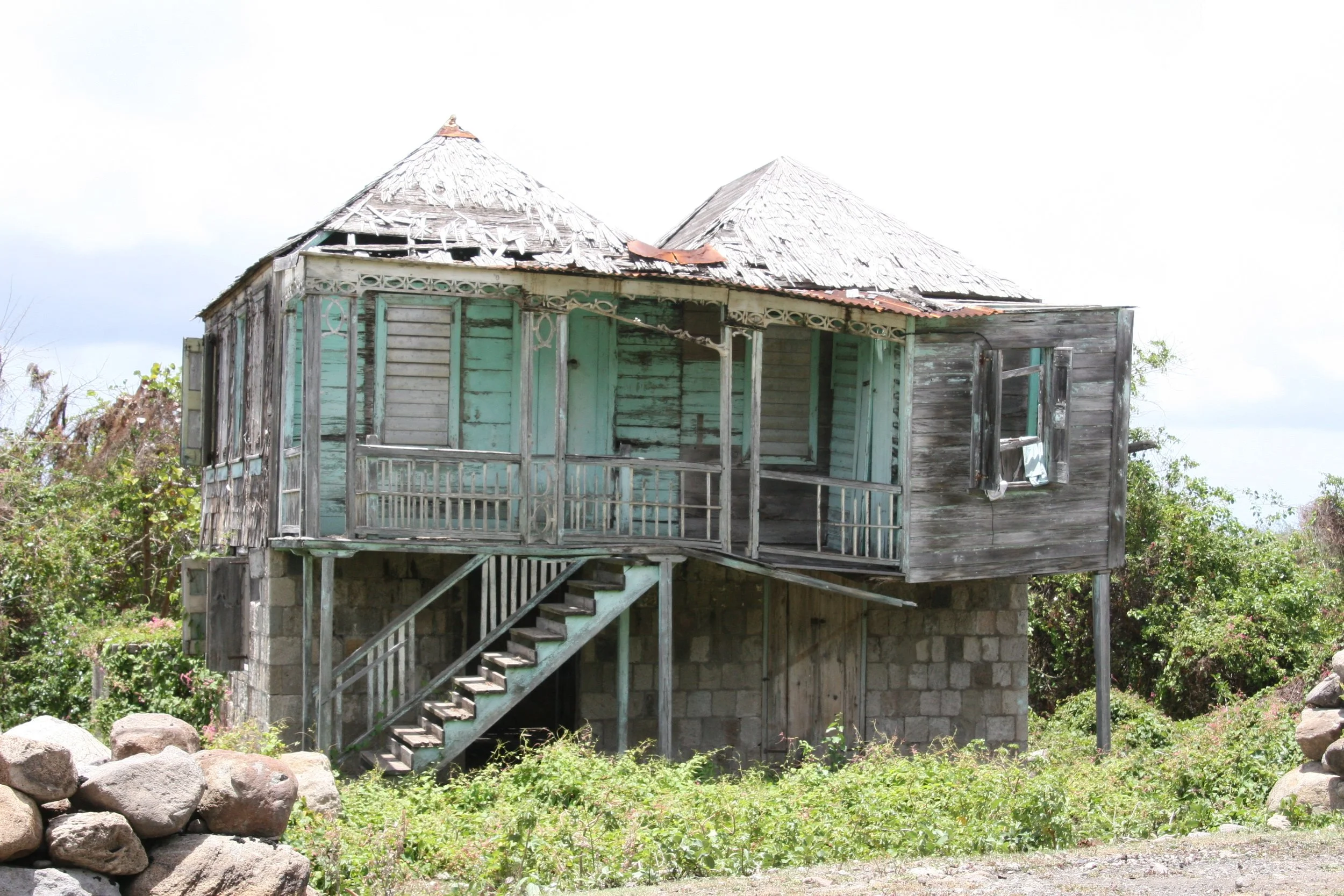 A weathered wooden house on stilts with a partially damaged roof, surrounded by greenery and a stone wall in the foreground.