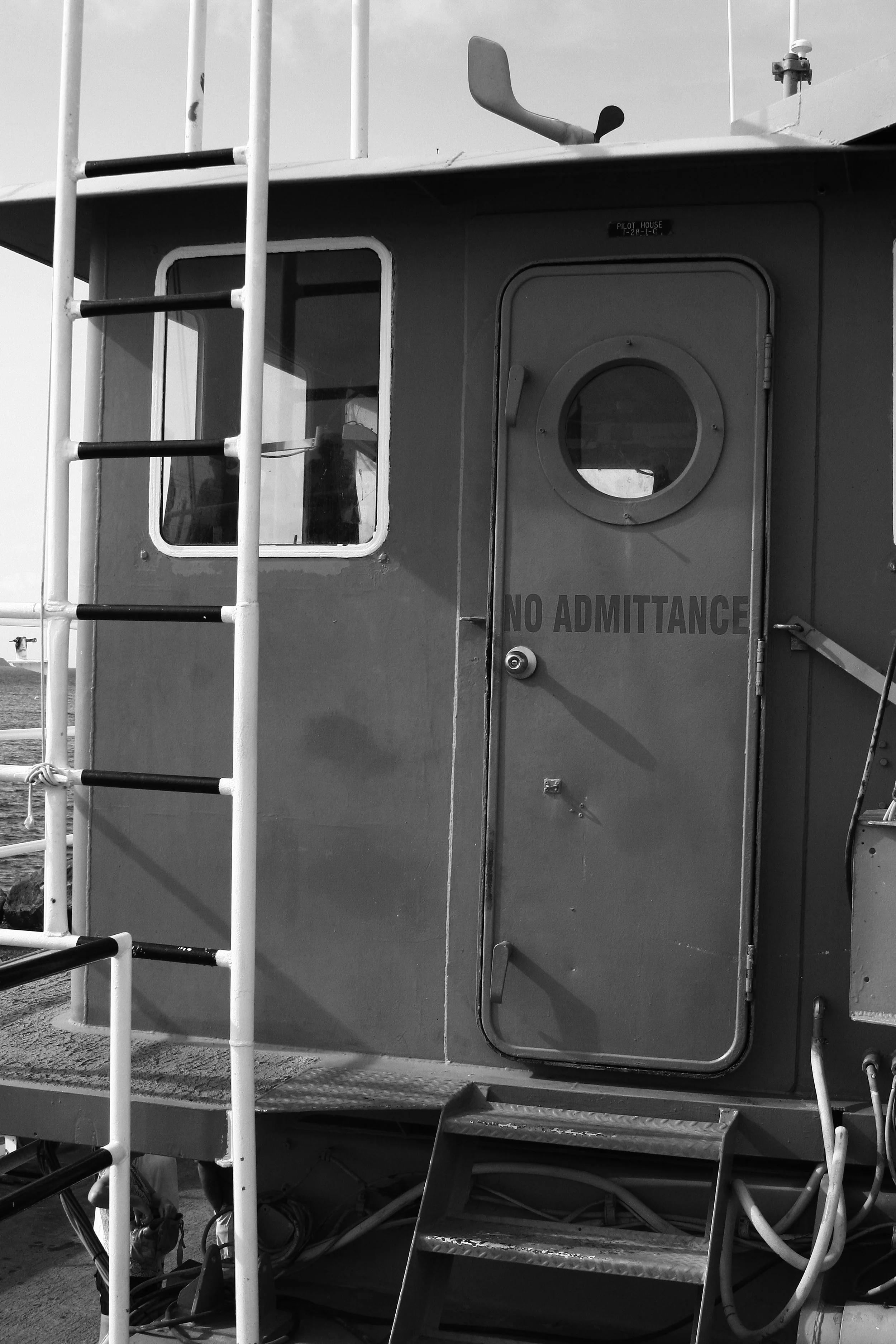 Black and white photo of a boat's cabin with the sign 'No Admittance' on the door, a window, and a ladder on the left side.
