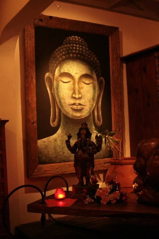 A dimly lit interior decor scene featuring a large framed painting of Buddha with closed eyes, a serene expression, and a textured head, mounted on a wall. In front of the painting, there is a small statue of a deity with multiple arms, a potted plan
