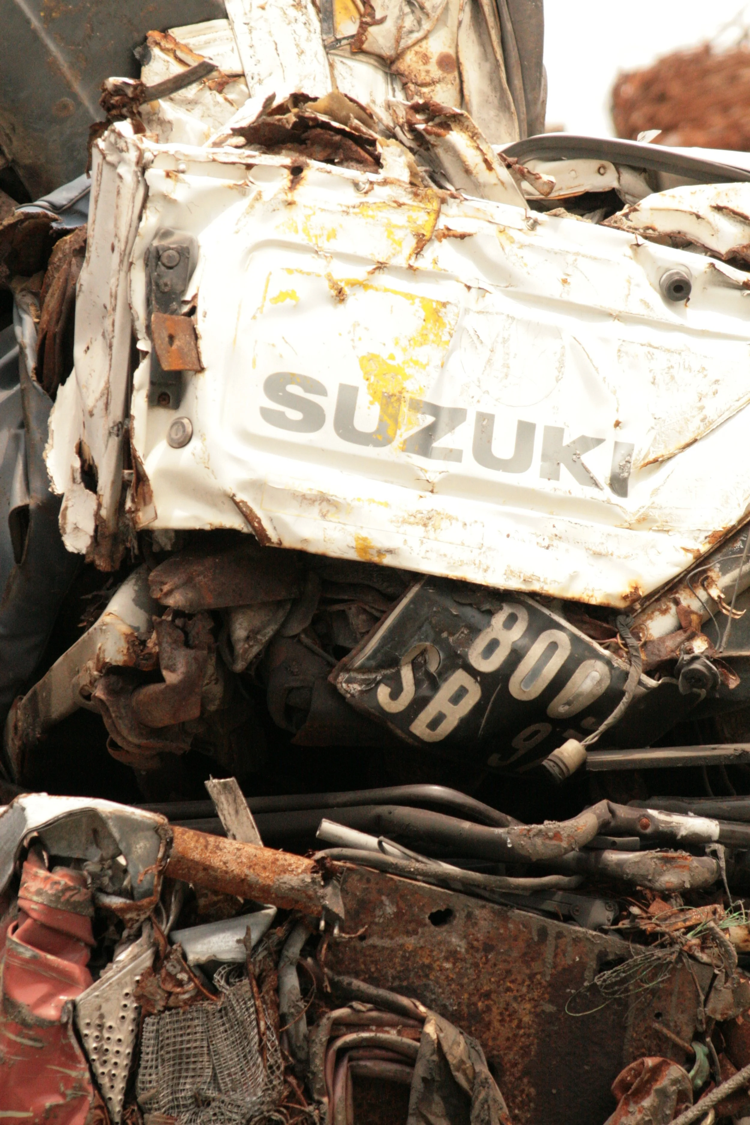 Close-up of a wrecked Suzuki vehicle with visible rust, dents, and damage to the body, including a damaged license plate.