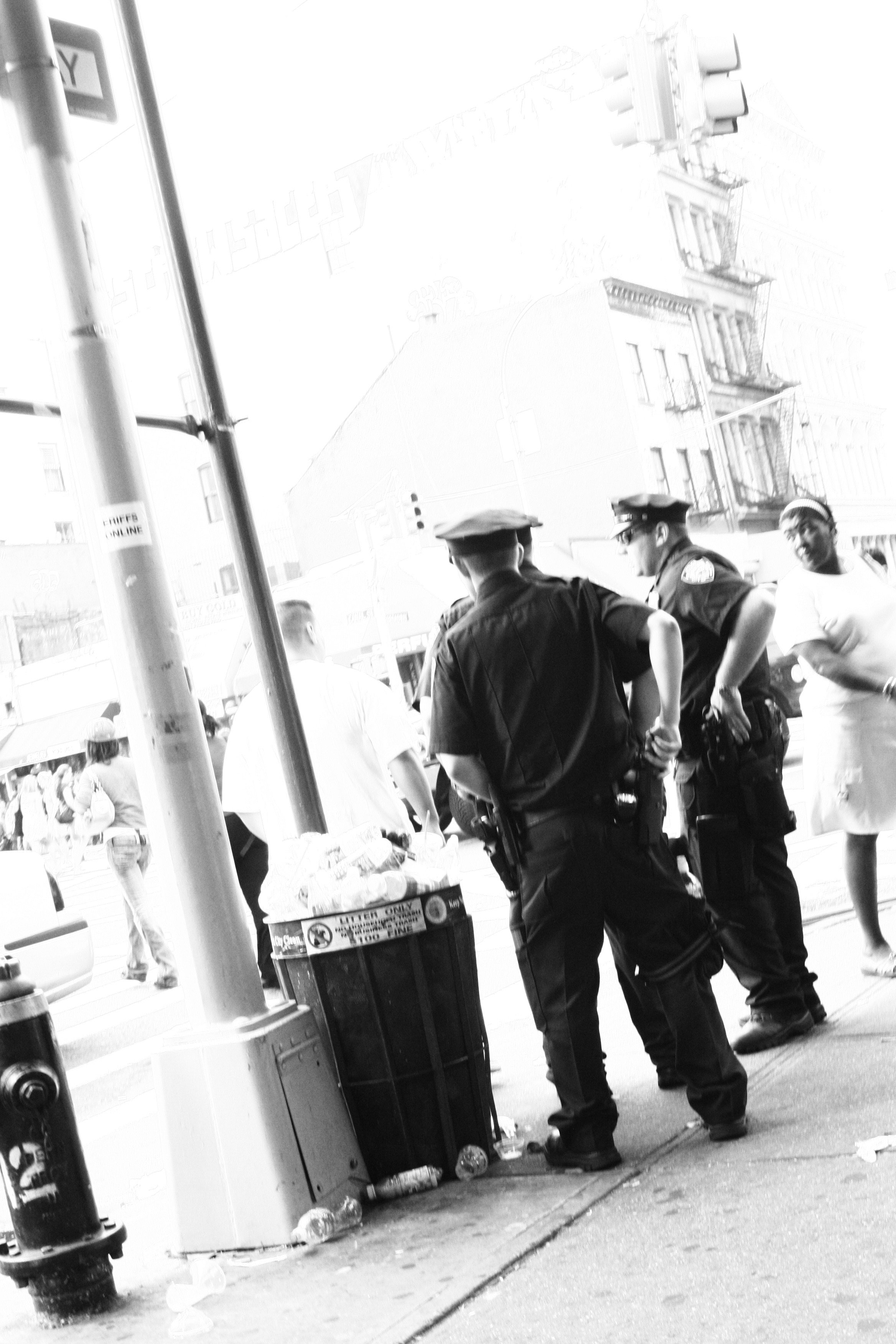 Two police officers standing on a city sidewalk, facing each other, talking. Around them are pedestrians, a trash can, and a street corner with buildings and traffic lights in the background.