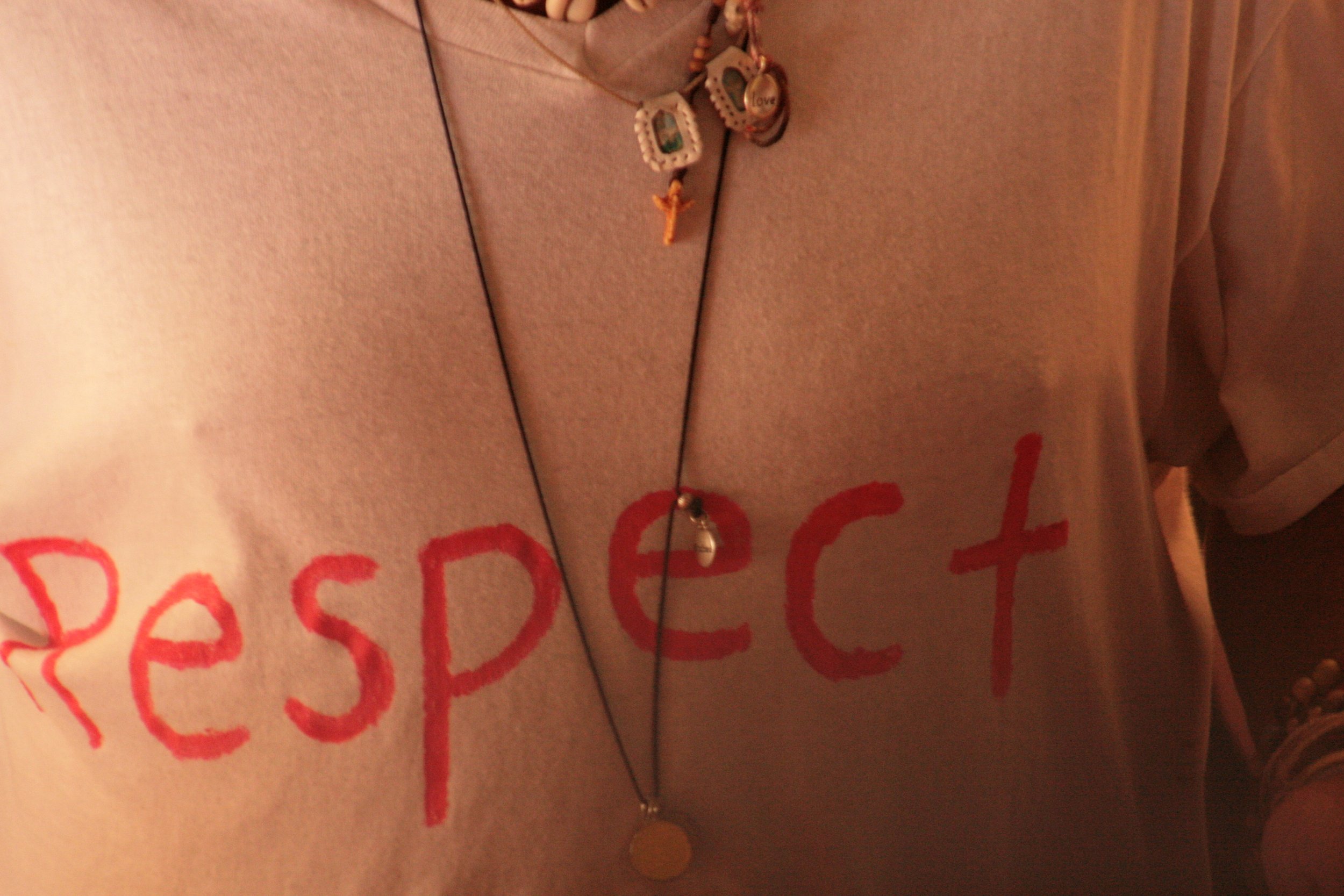 Close-up photo of a shirt with the word 'RESPECT' written in red on it, and layered necklaces, one with a cross pendant and another with a round pendent.