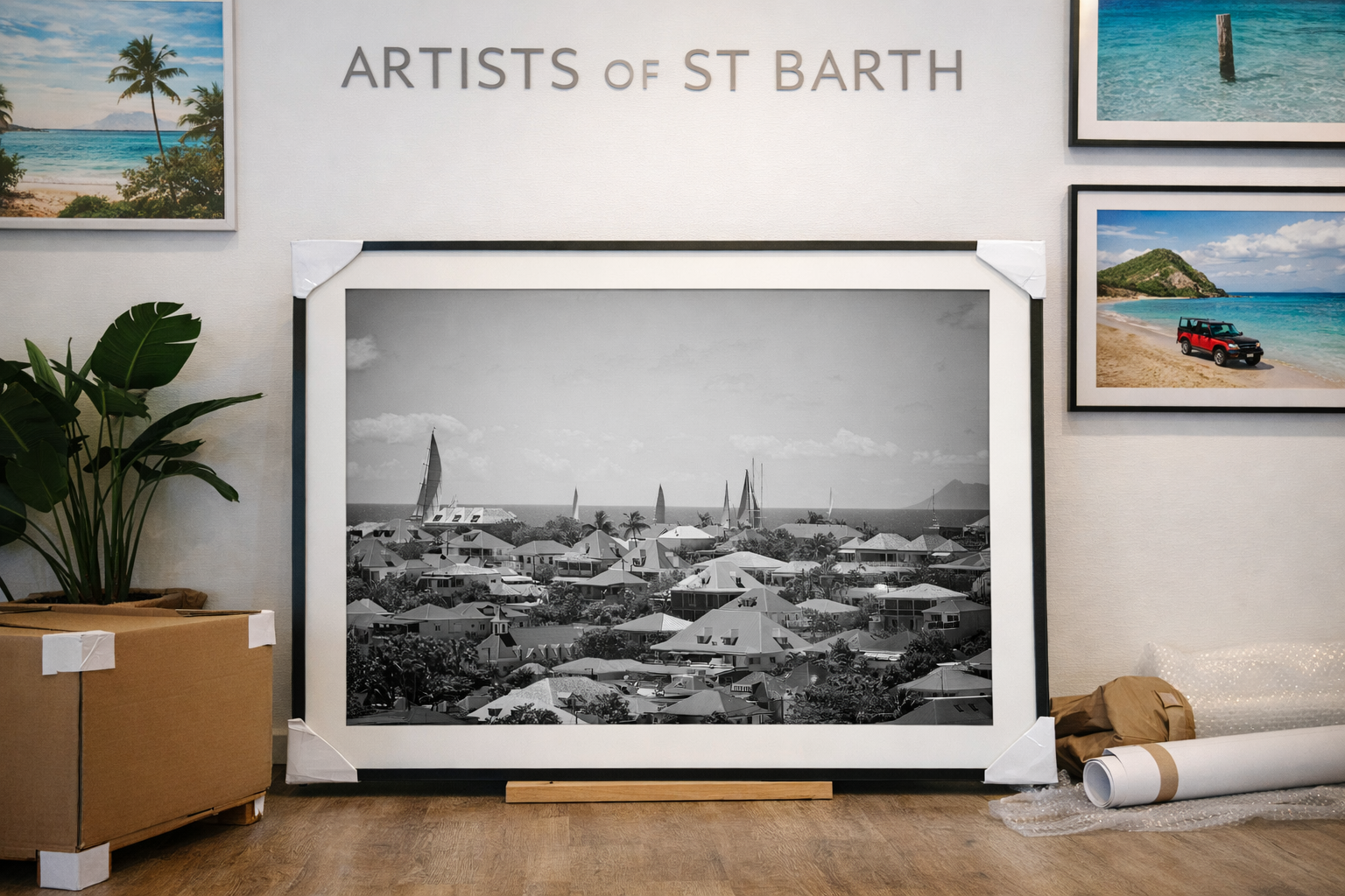A framed black and white photograph of a coastal town with sailboats in the water, set on a wooden floor against a wall. There are framed photographs of beach scenes and an indoor plant nearby, with packing materials on the floor.