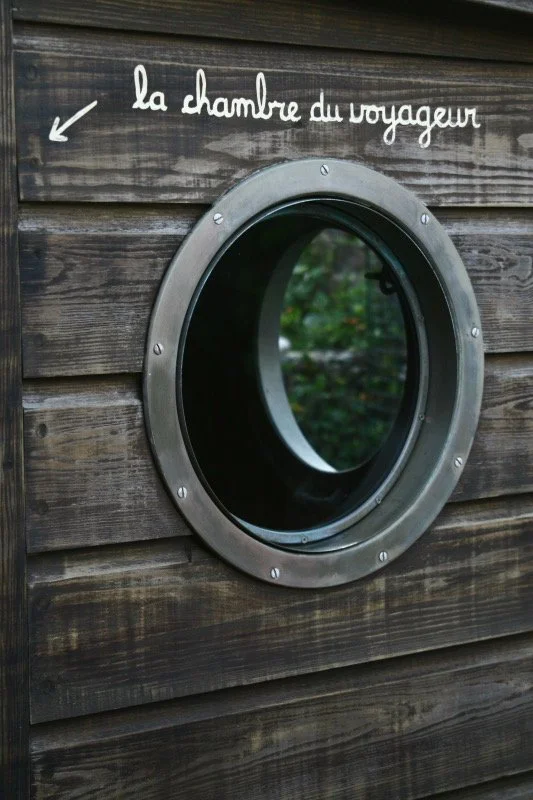 A round window with a metal frame built into a wooden wall, with a sign above it that says 'la chambre du voyageur' and an arrow pointing to the window.