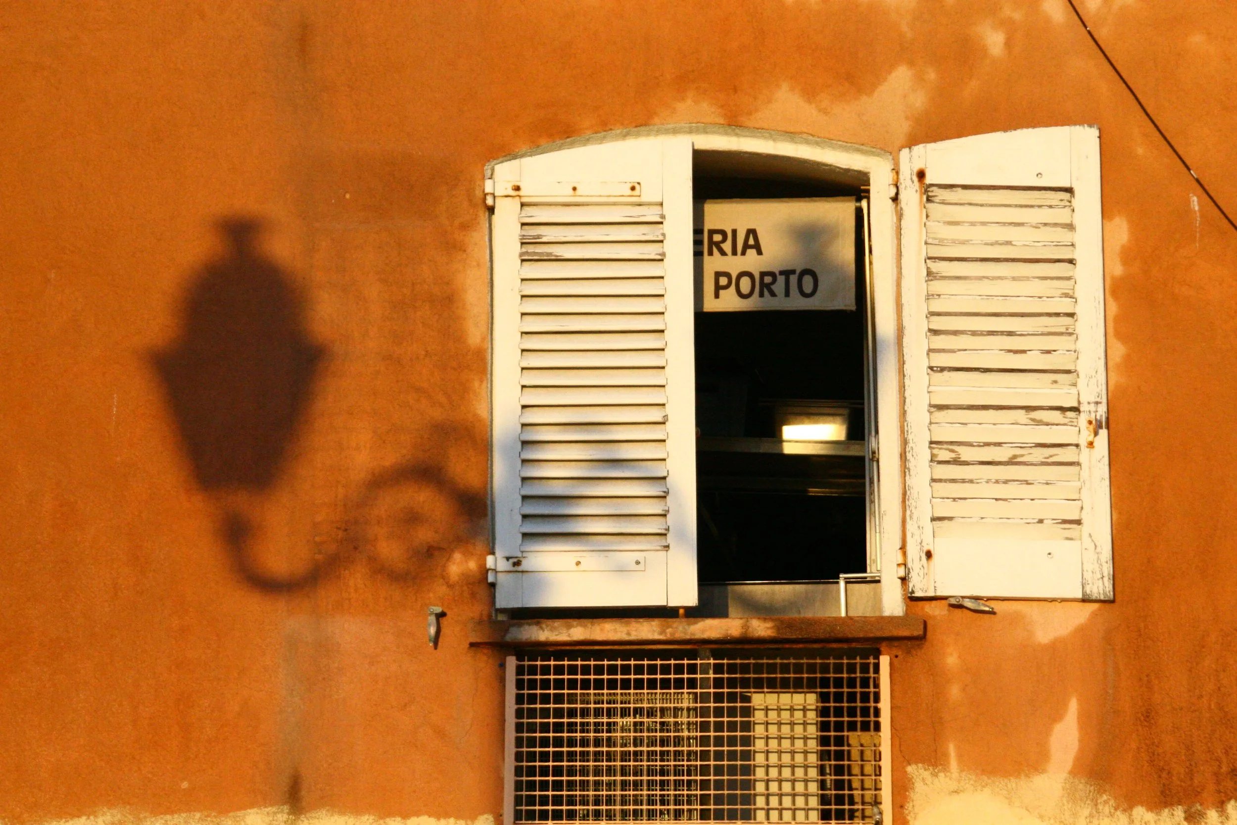 A warm-colored wall with a small window and open shutters casting a shadow of a bicycle and a person. The window has a sign that reads 'RIA PORTO'.