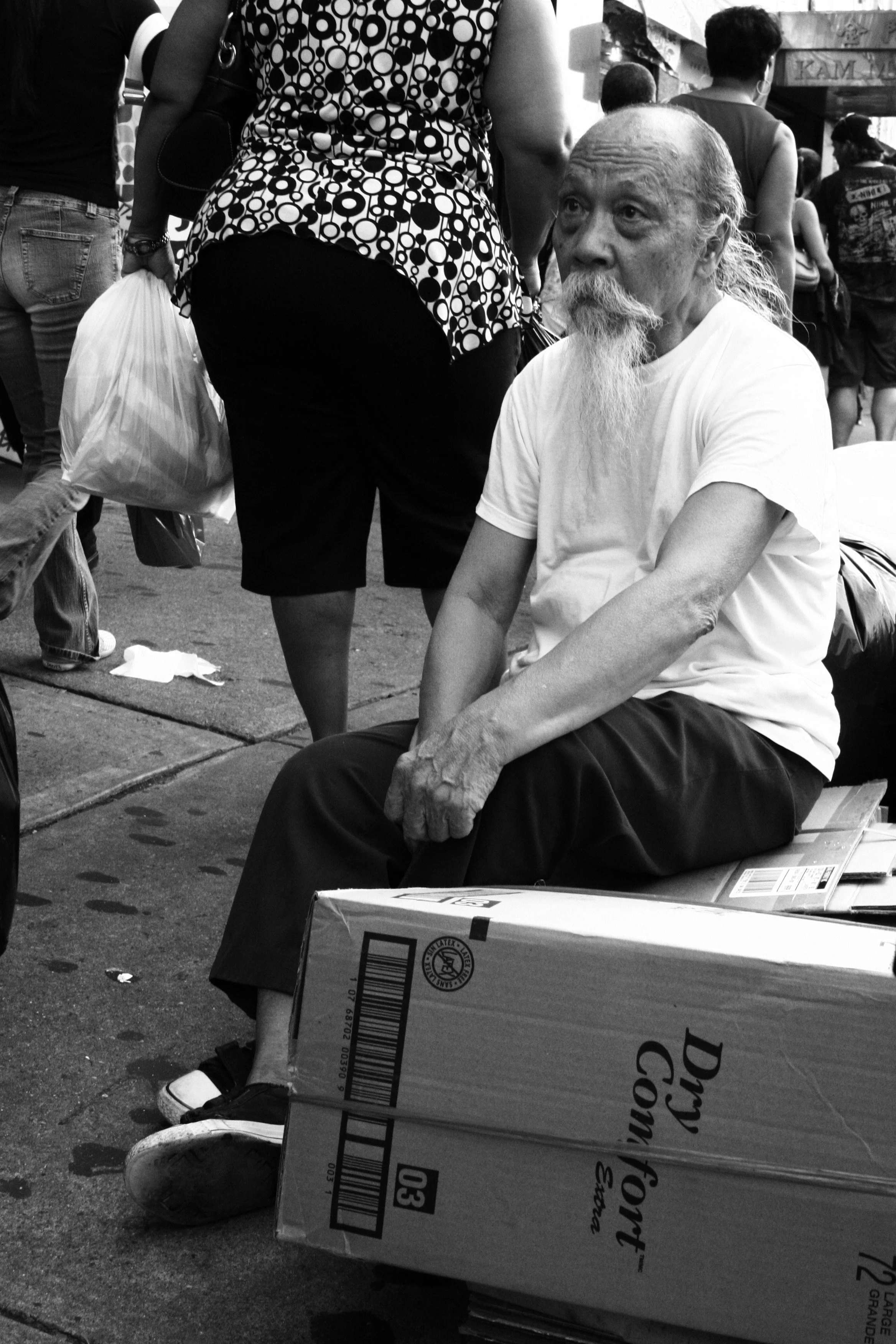 An elderly man with a long white beard sitting on a box on a crowded city street, wearing a white T-shirt and dark pants.