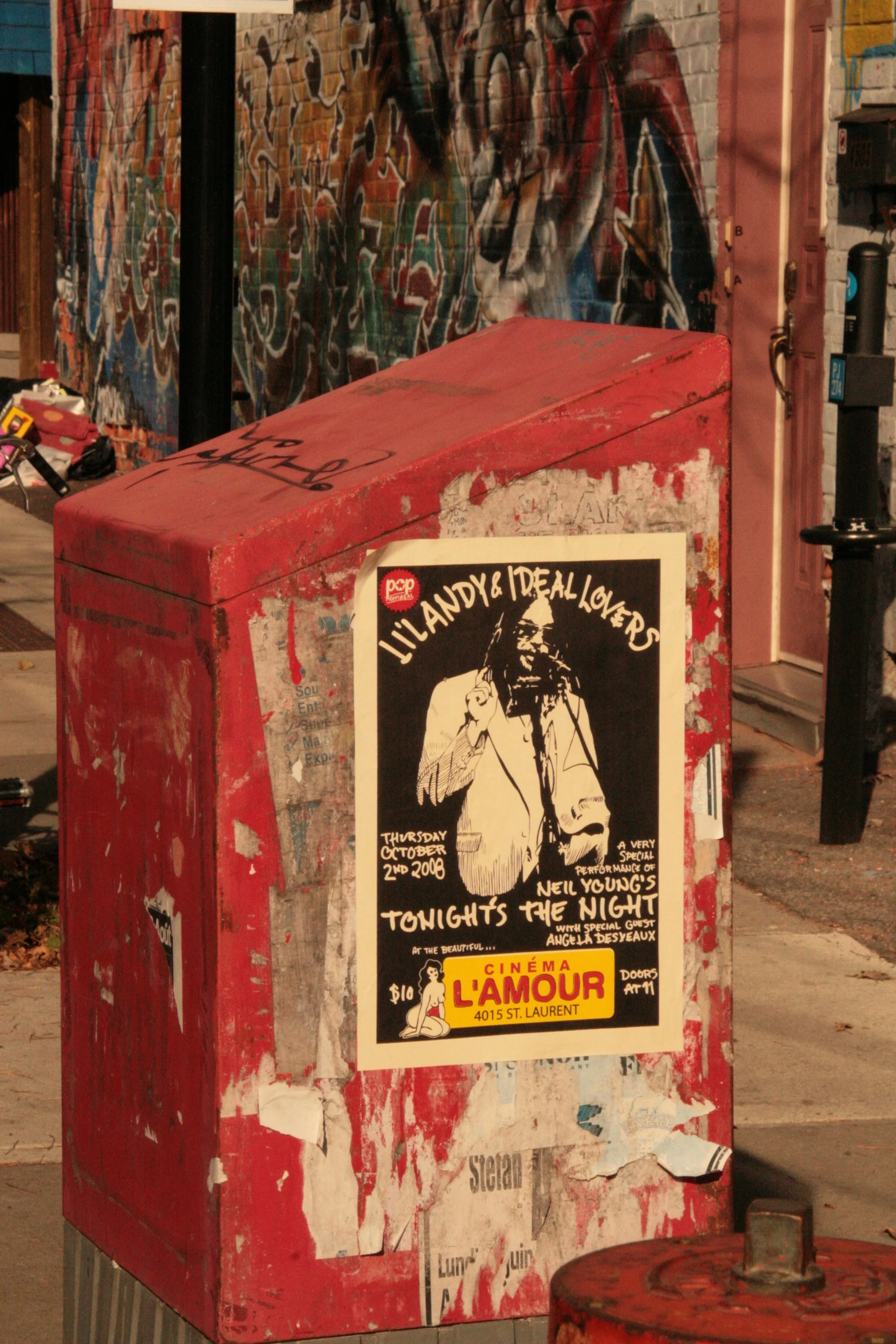 A red, weathered newspaper box with a poster advertising an event at Cinéma L'Amour at 4015 St. Laurent. The poster features a black and white drawing of a man with dreadlocks and glasses, holding a microphone, and includes details of an October 2nd,