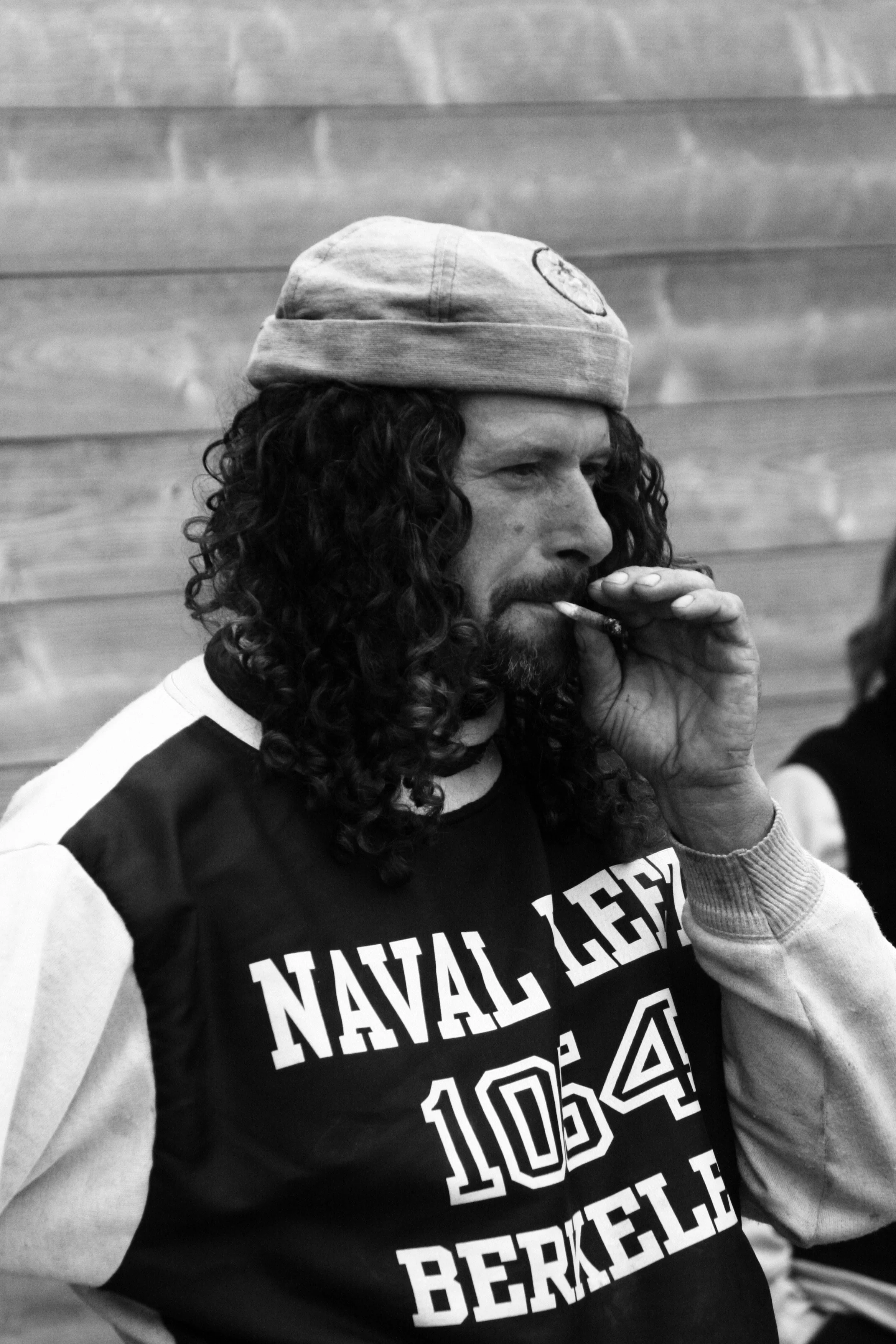 A man with long curly hair, wearing a beanie and a black and white sweatshirt that reads 'NAVAL ACADEMY 104 Berkeley,' is smoking a cigarette.