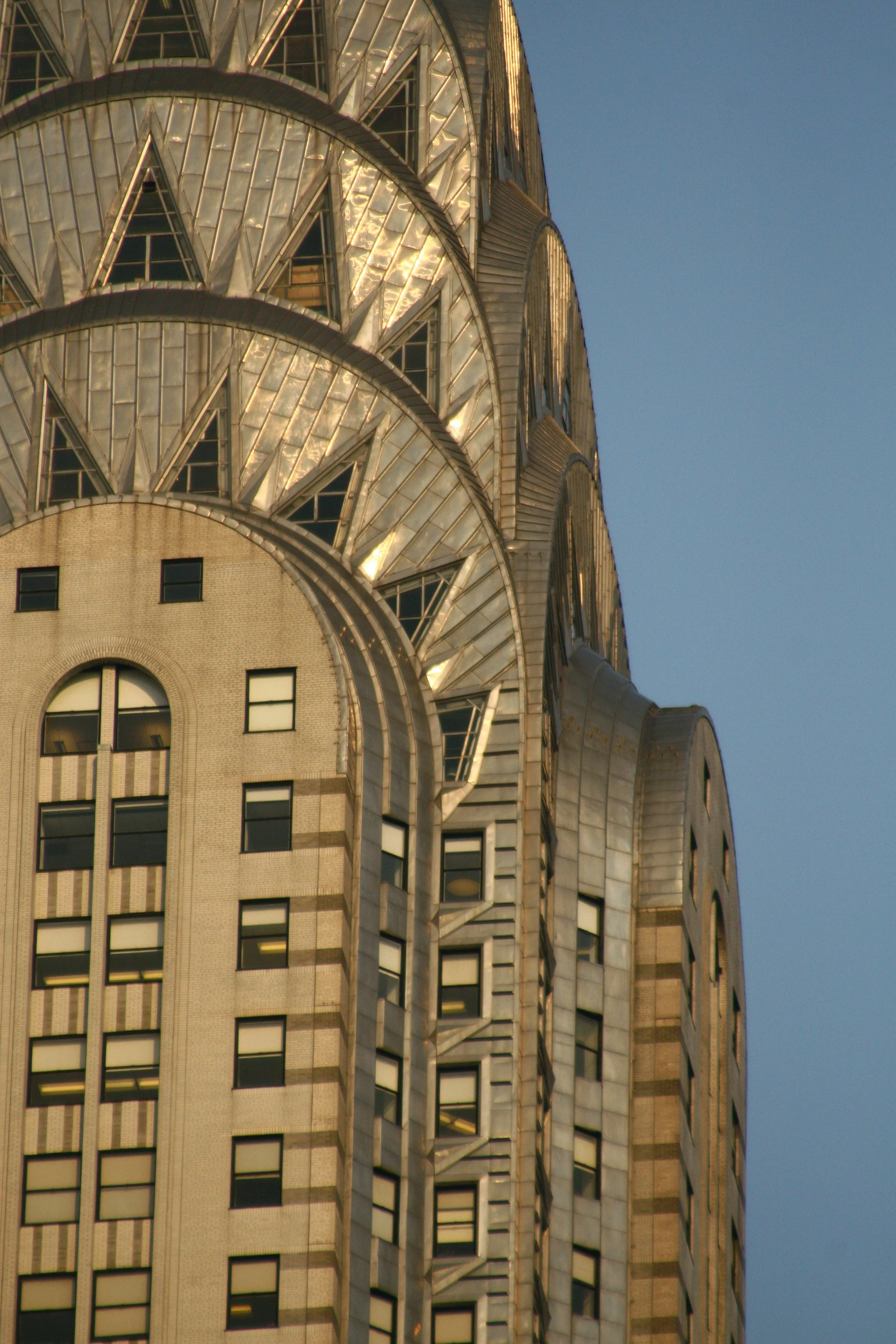 Close-up of the top section of the Chrysler Building in New York City, featuring its distinctive Art Deco architecture with terraced crown and triangular windows against a clear blue sky.