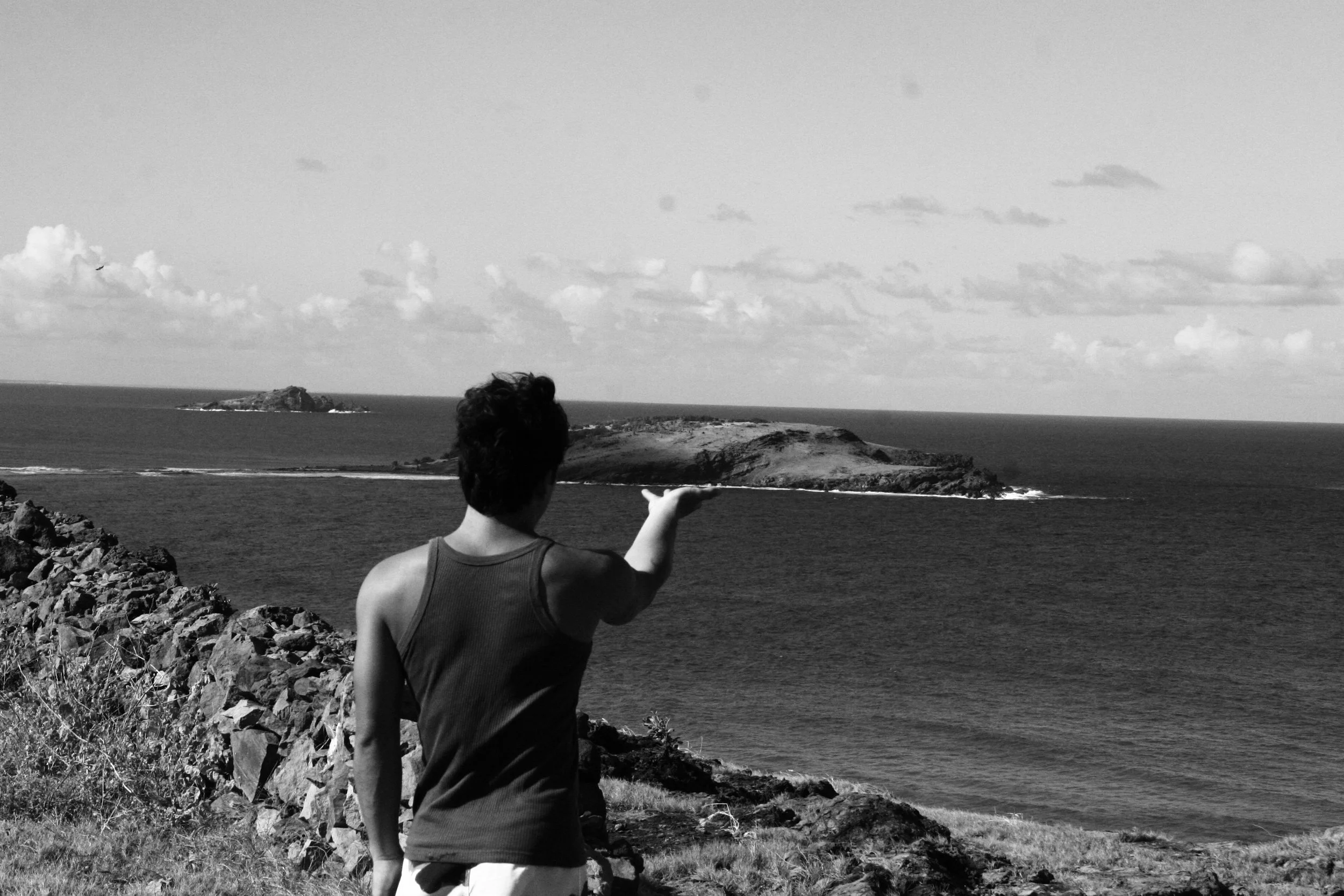 A person standing on rocky terrain pointing towards an island across the ocean in a black-and-white photo.