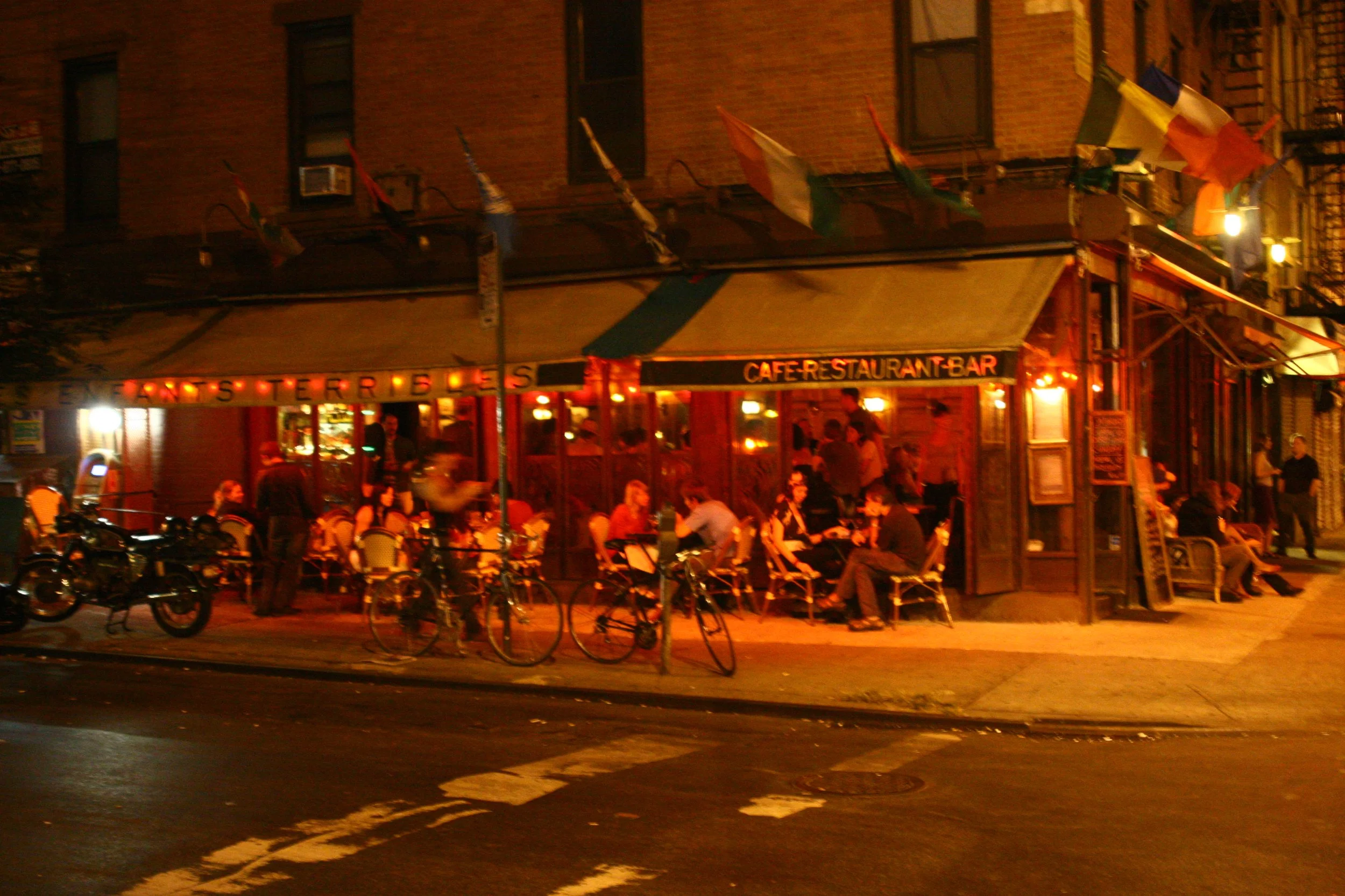 Nighttime scene of an outdoor cafe with people sitting on chairs and bicycles parked outside. The cafe has a yellowish awning and surrounding warm lighting, with signage indicating it is a restaurant and bar.