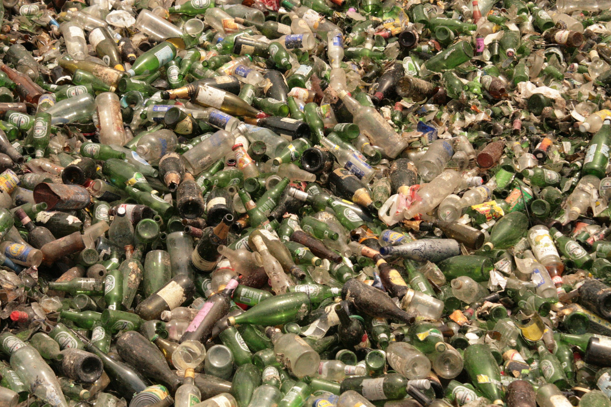 A large pile of broken glass bottles, mostly green glass beer bottles, some with labels. The bottles are dirty and discarded in a junkyard or recycling center.