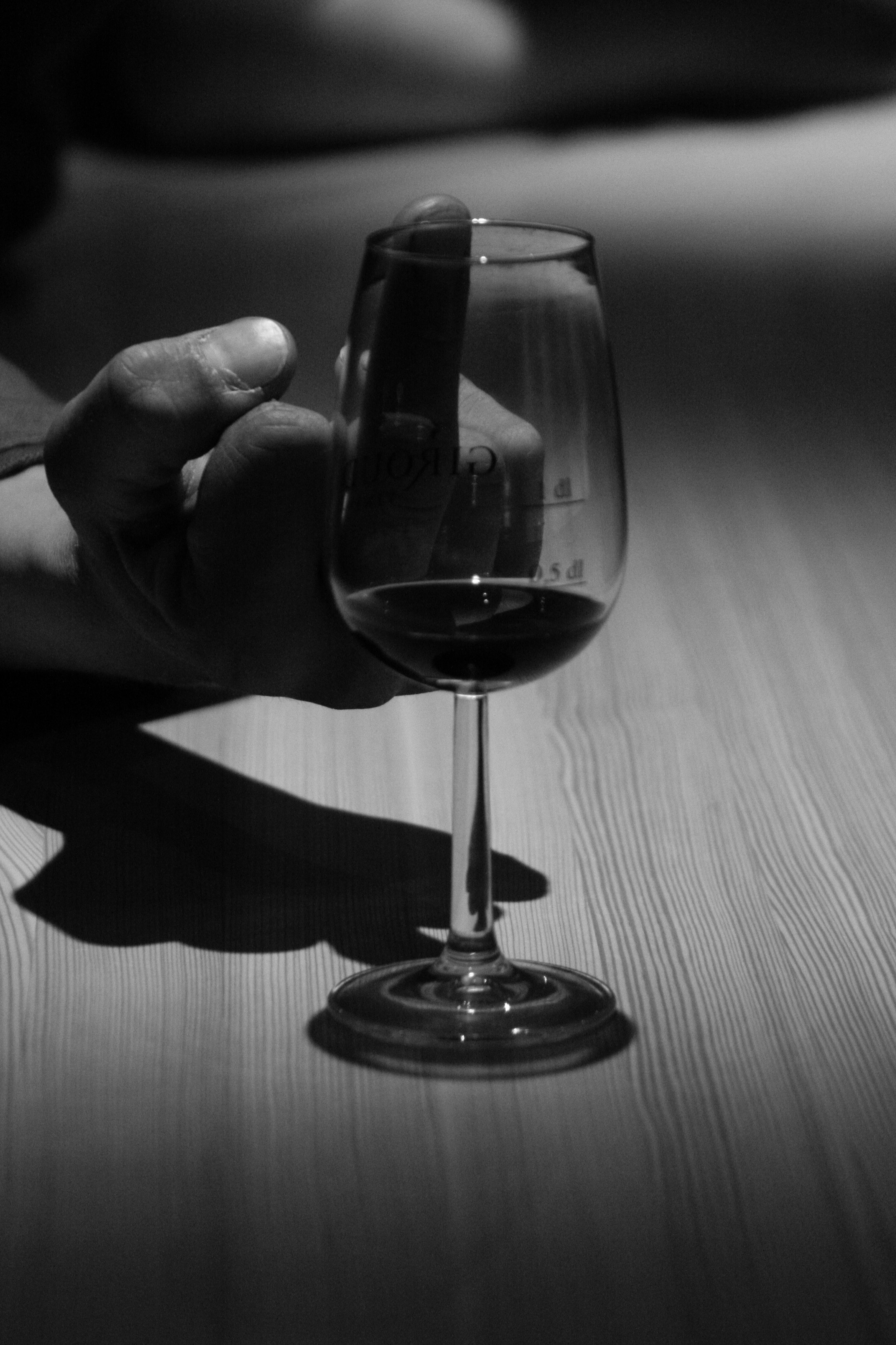 Black and white photo of a hand holding a wine glass filled with red wine, casting a shadow on a wooden table.