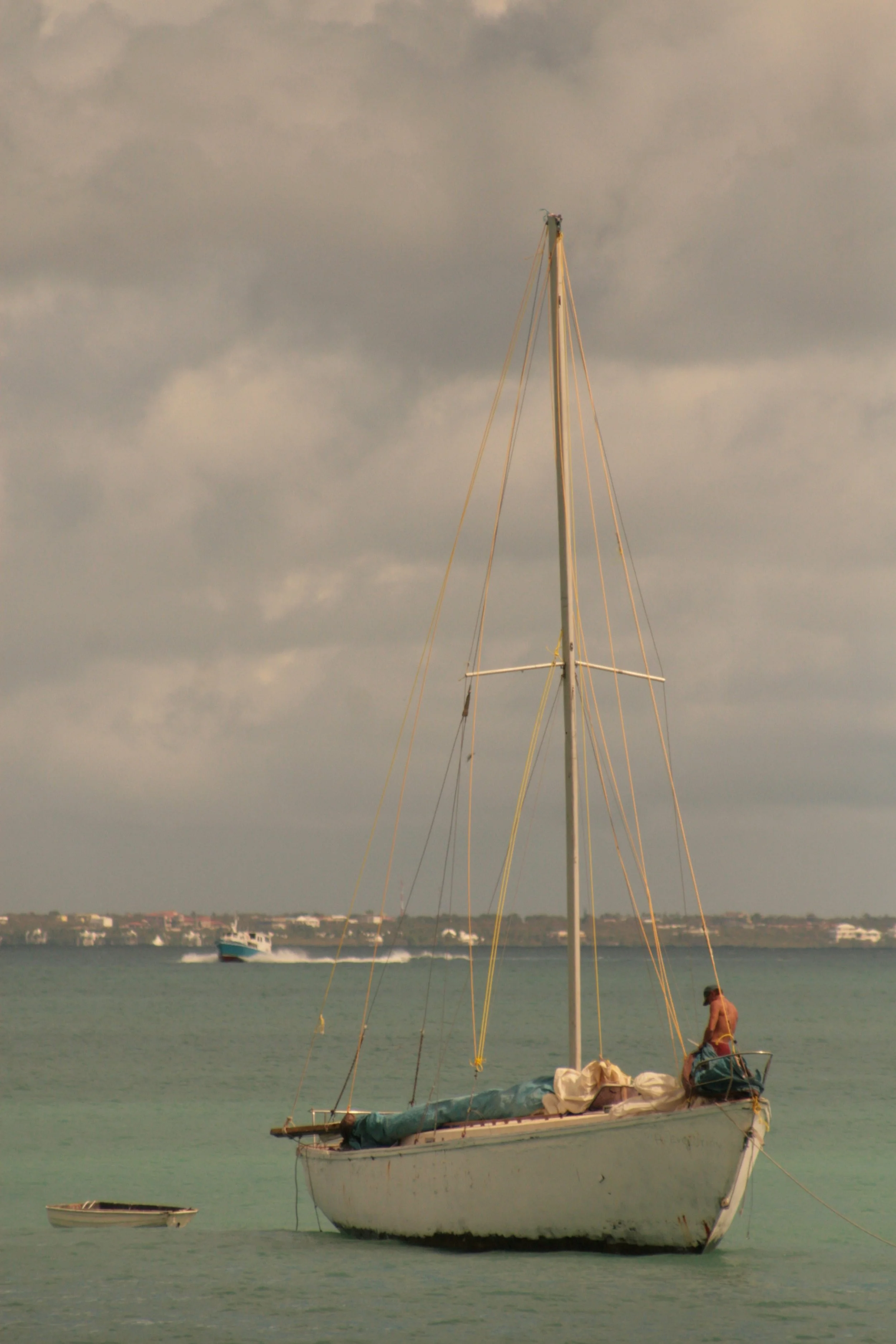 A white sailboat with a person on deck, docked on calm water during cloudy weather. A smaller boat is floating nearby. In the background, there is a distant shoreline with buildings.