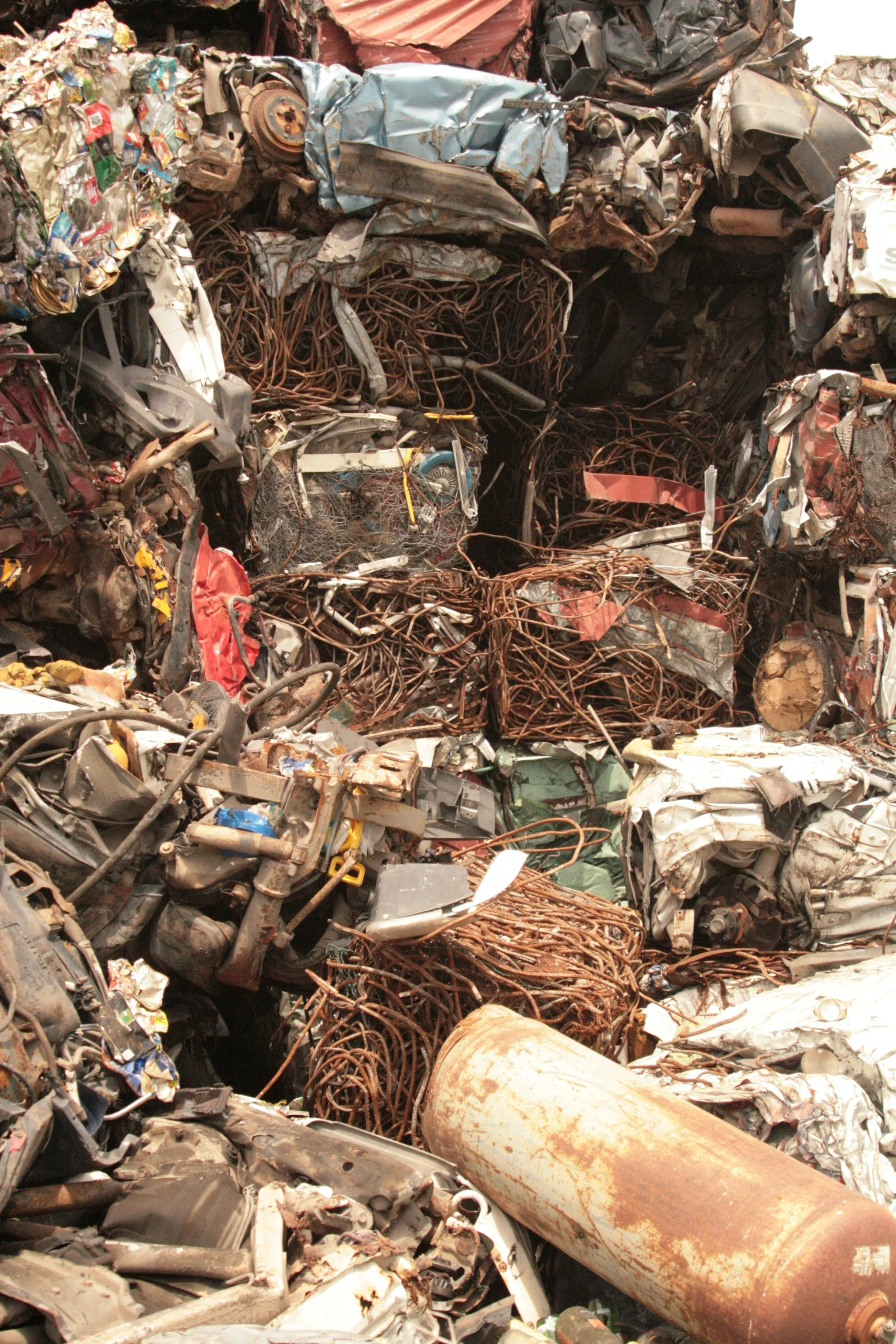 A pile of crushed and scrap metal, including twisted wires, old cars, and rusty cylindrical objects, in a junkyard.