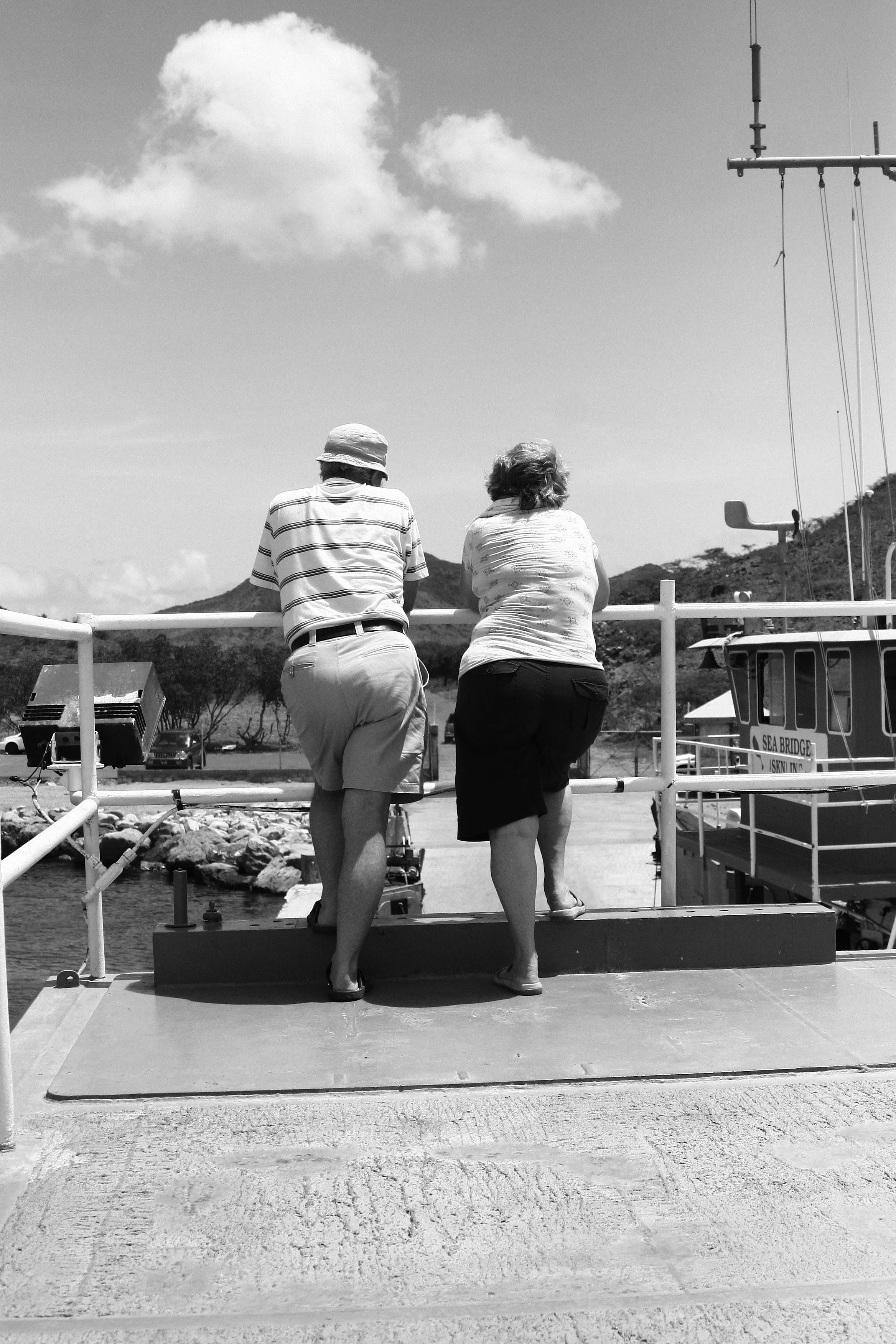 Two elderly people, a man wearing a hat and striped shirt and a woman wearing a patterned top, are standing near the edge of a dock, looking out over the water with boats and hills in the background.