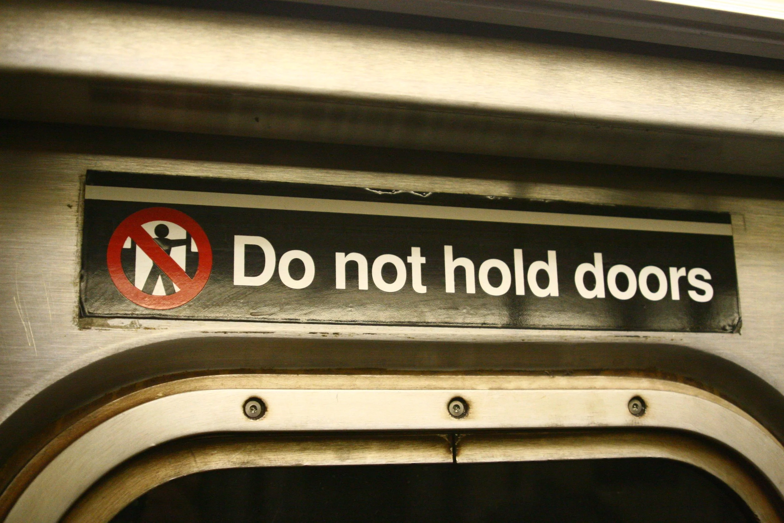 A sign on a train door that says 'Do not hold doors' with a pictogram of a person holding a door, crossed out in red.