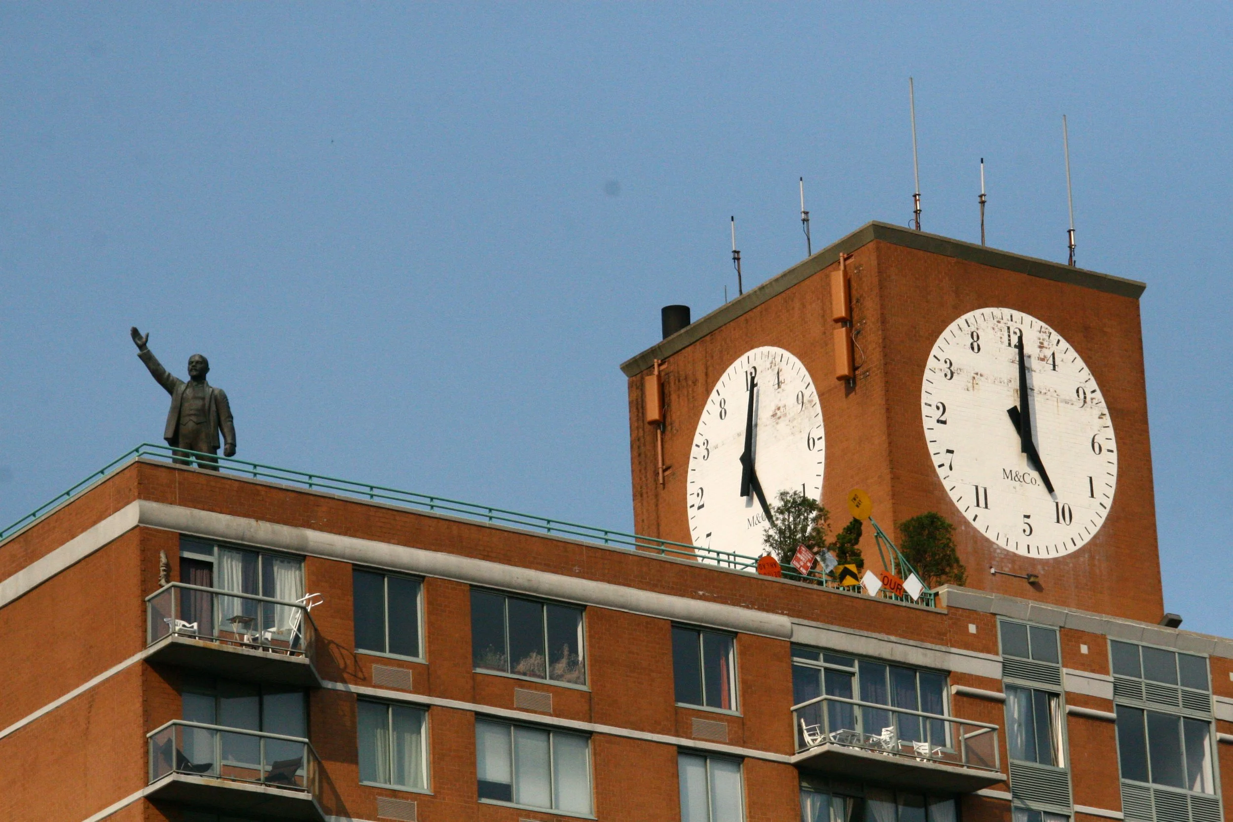 A brick building with multiple windows and small balconies, with a clock tower showing approximately 12:01, and a statue of a man waving on the roof.