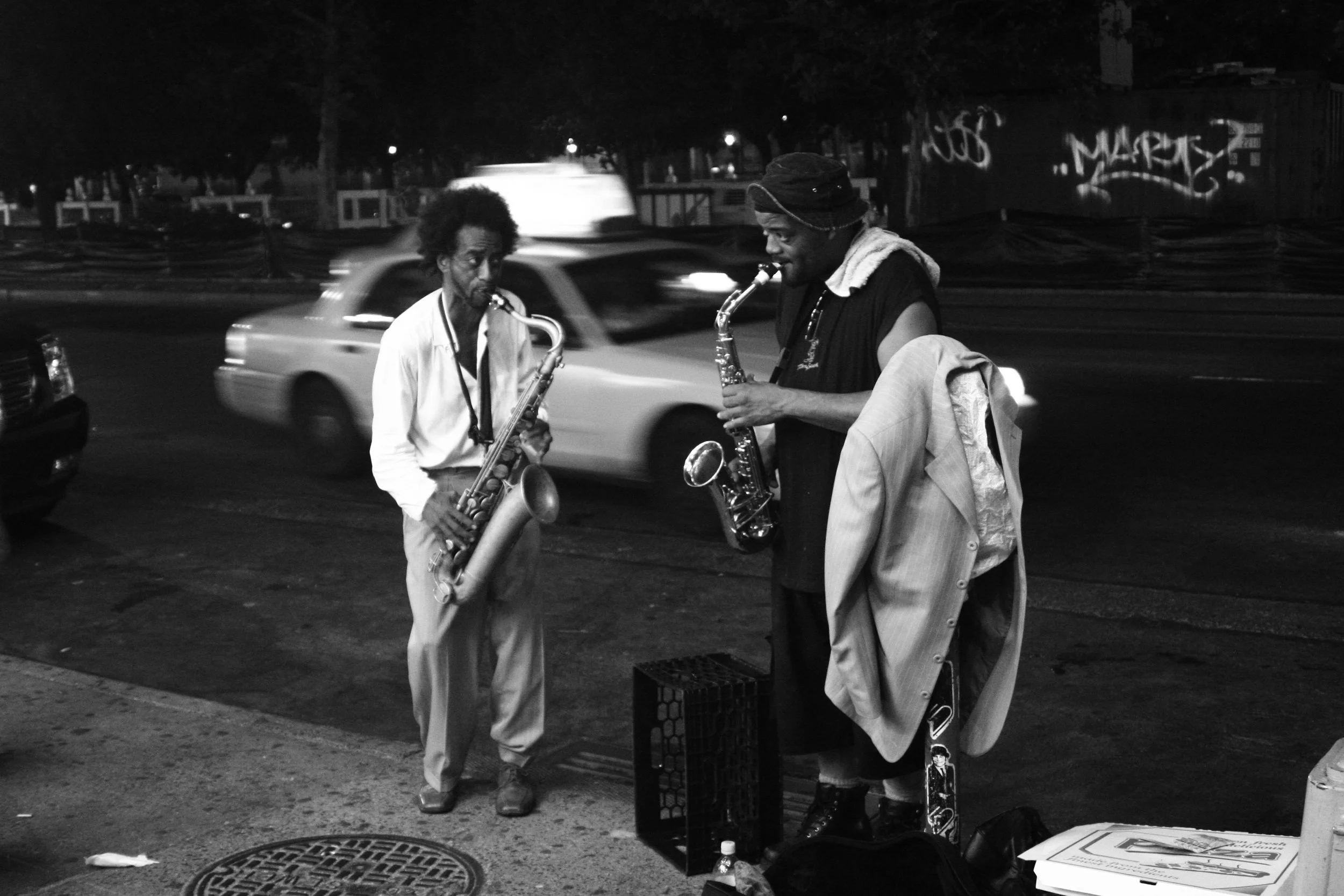 Two street musicians playing saxophones on a city sidewalk at night, with cars passing by in the background in black and white.