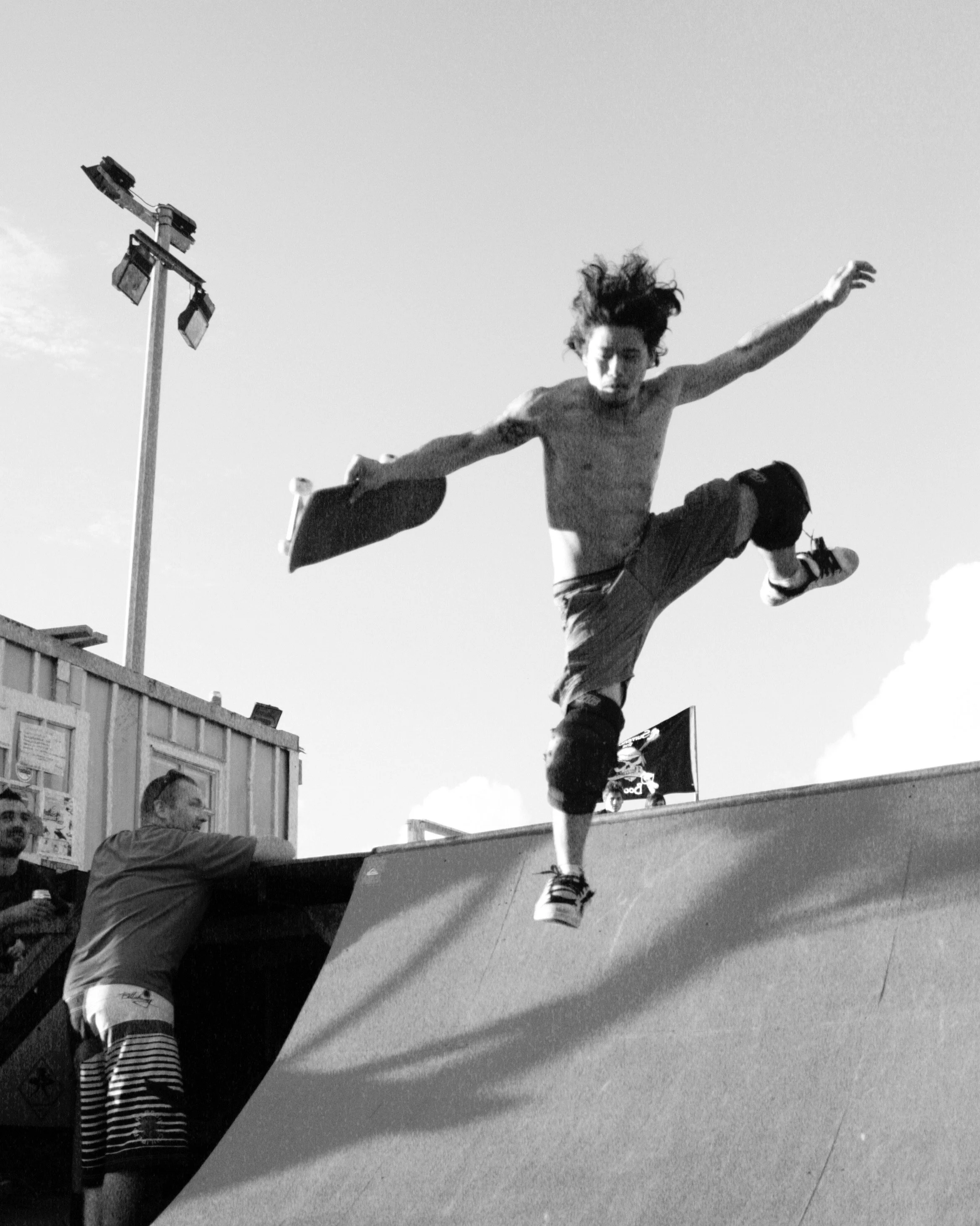 A shirtless skateboarder with long hair is mid-air at a skate park, executing a jump or trick, while a couple of spectators watch nearby.