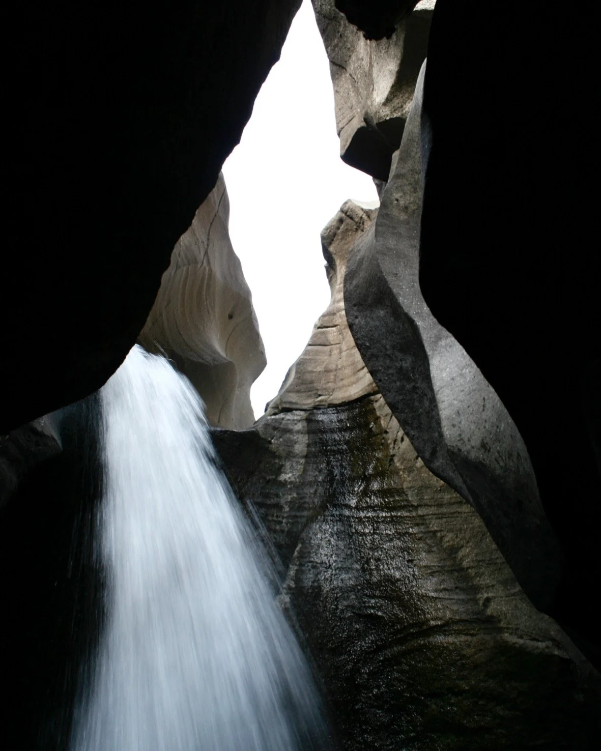 Waterfall flowing through a narrow canyon between large, dark rocks with a bright sky visible at the top.